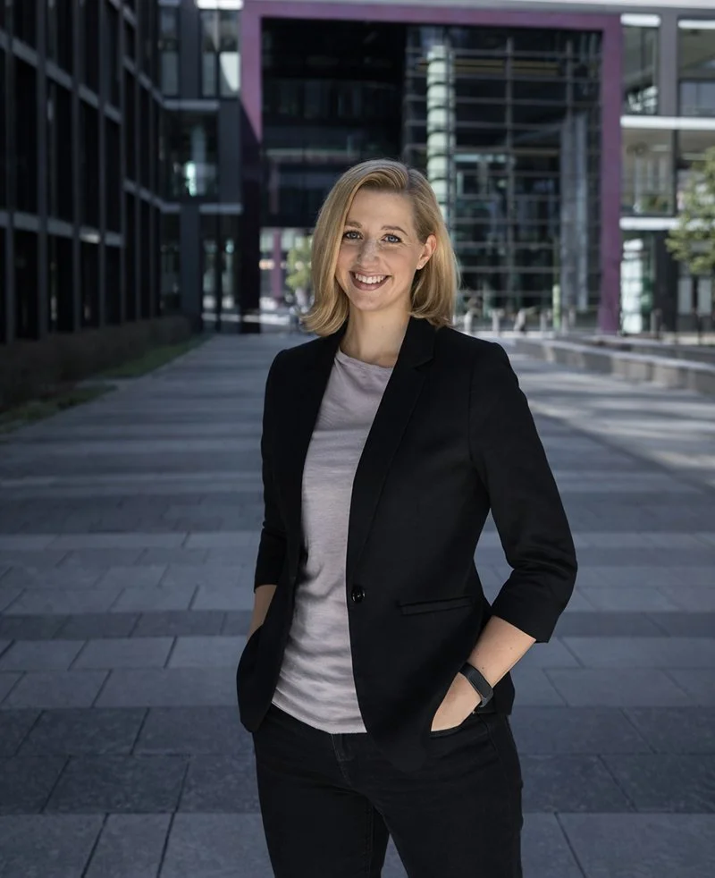 Frau in schwarzem Blazer und grauem T-Shirt, lächelnd, steht vor moderner Gebäudefassade, Außenaufnahme.
