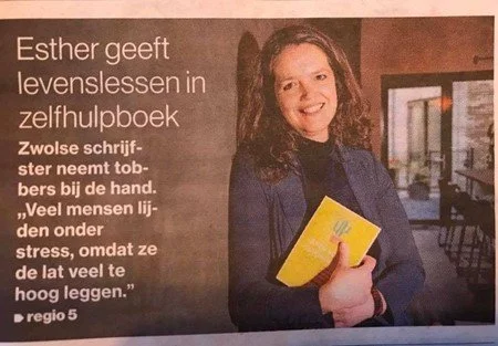 Een vrouw met bruin haar, gekleed in een donkerblauwe jas, houdt een geel boekje vast en lacht naar de camera. Op de achtergrond is een raam en binnen een stoel zichtbaar.