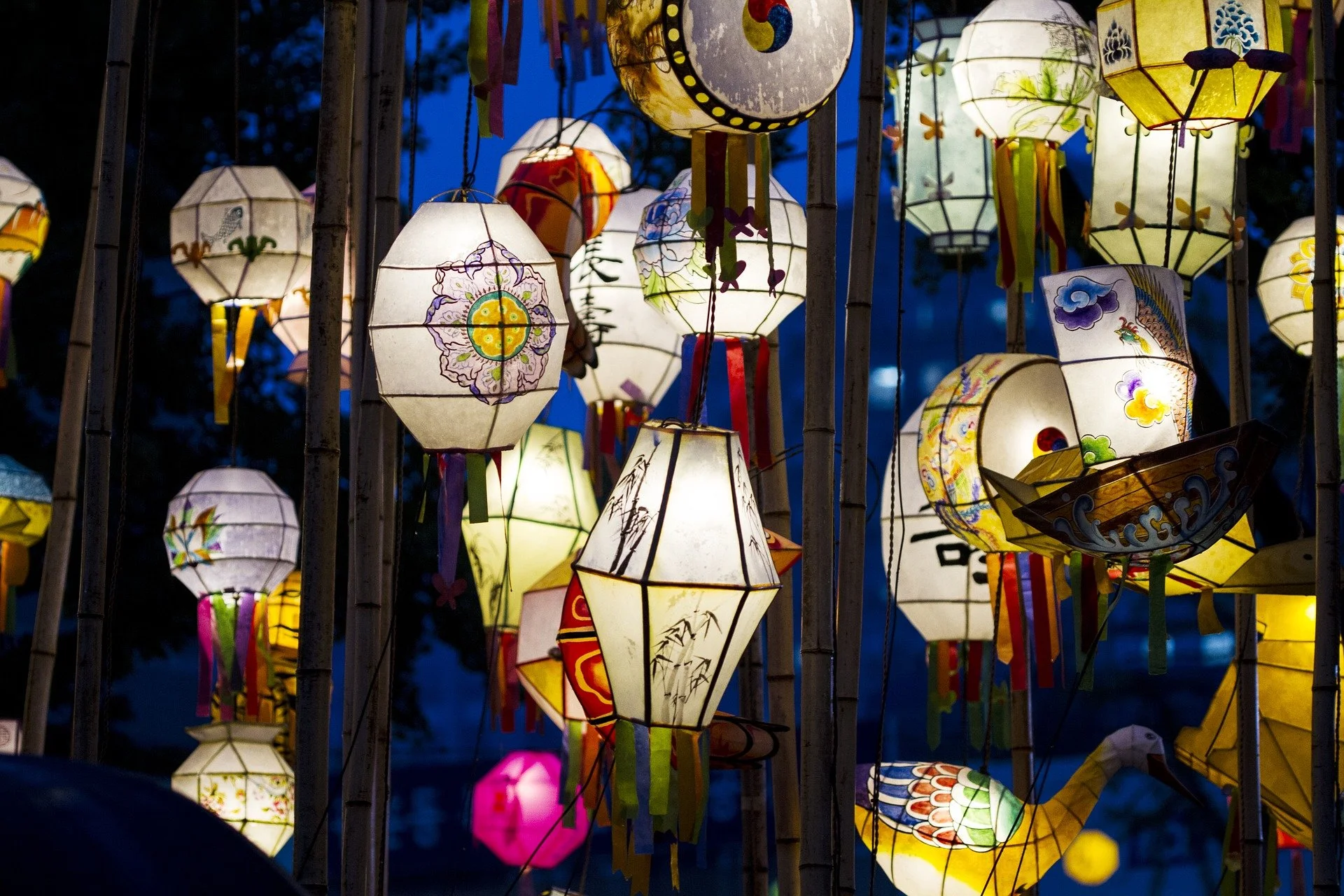 Verspreide kleurrijke paper lanterns hangen aan touwen in de avond, geven zacht licht, versierd met bloem- en diermotieven. Achtergrond is donkerblauw met silhouetten van bomen.