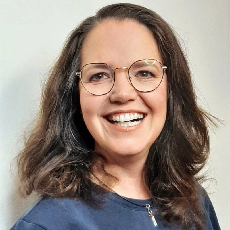 Een lachende vrouw met bruin, golvend haar en bril, gekleed in een donkerblauwe top.