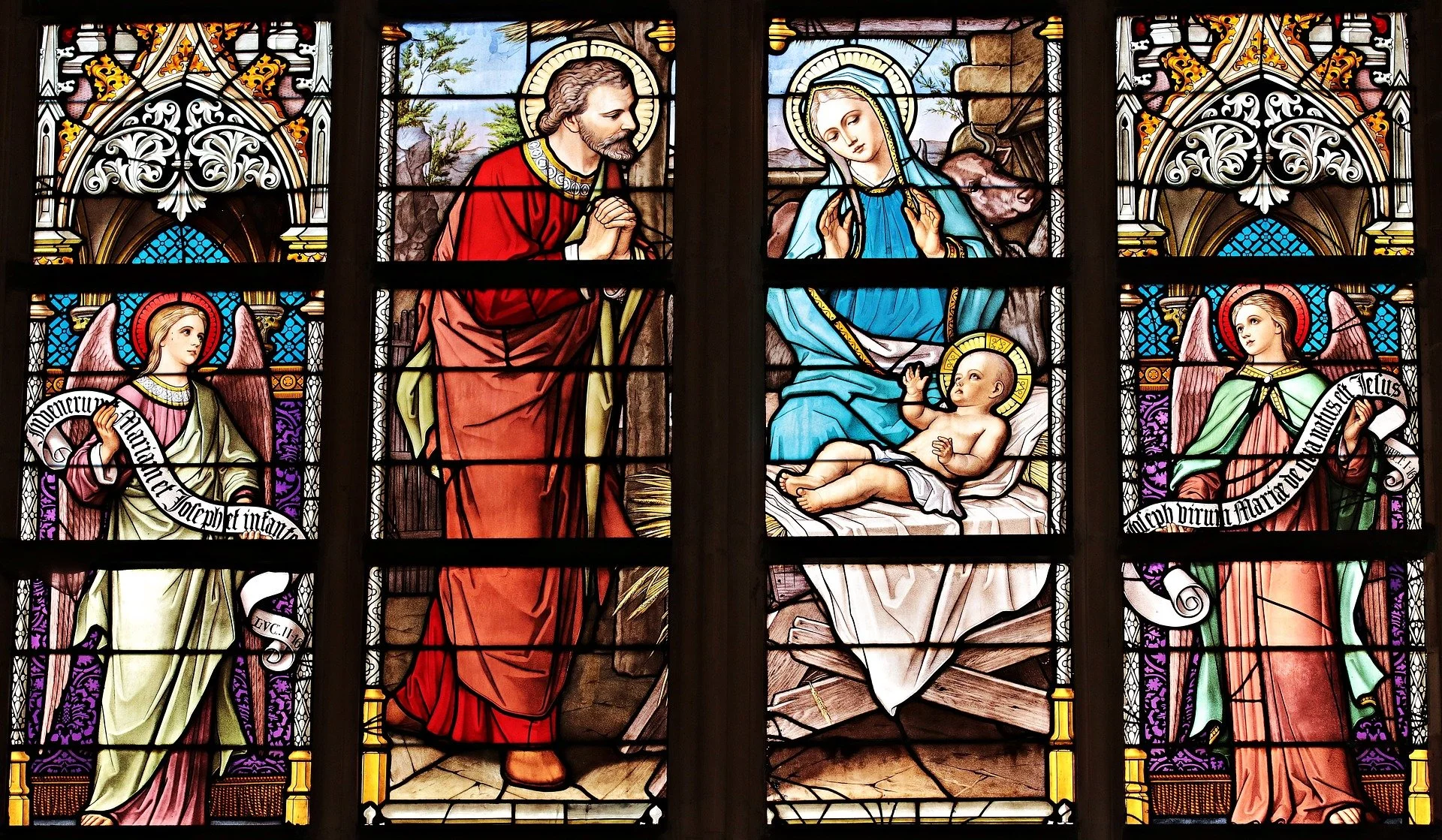 Kunstglas-in-lood raampanel met een religieuze scène van Maria met baby Jezus, geflankeerd door engelen, en een man die zich voor hen neerknielt.