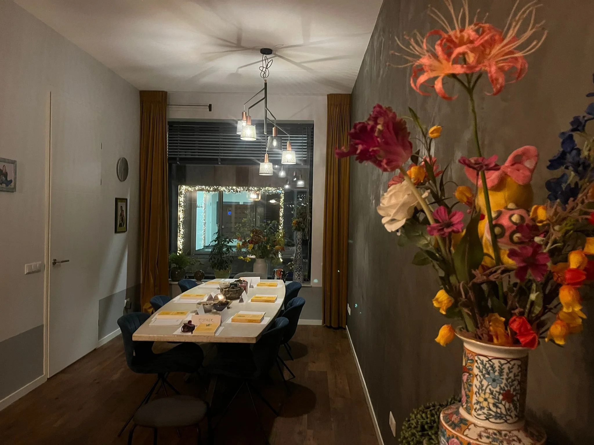 Een eetkamer met een grote tafel, omringd door acht stoelen, en een verzameling boeken op de tafel. In de achtergrond hangen moderne lampen en een grote raam met gordijnen. In een vaas rechts op de voorgrond zitten kleurrijke bloemen.