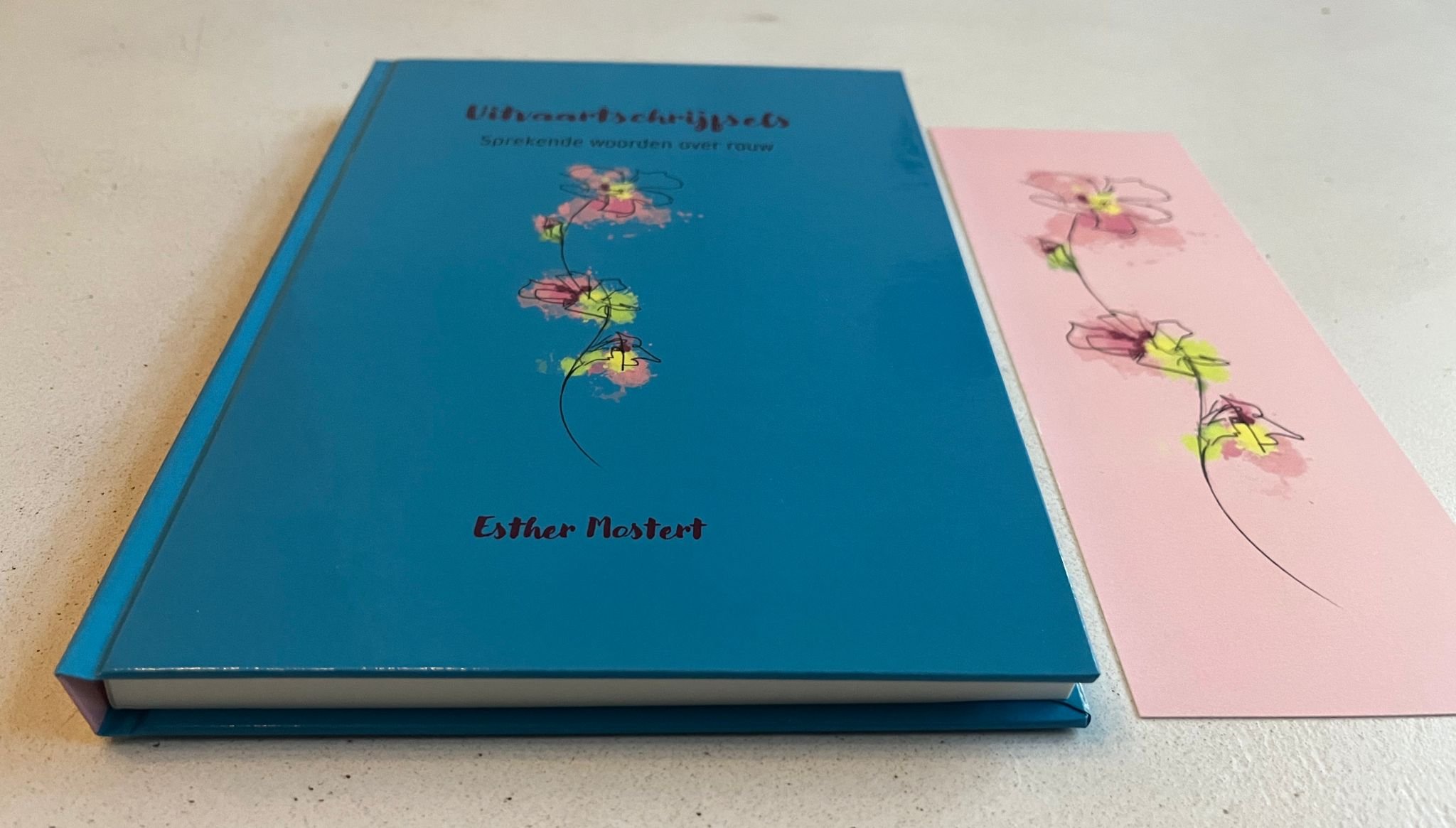 Twee boeken en een blad met een bloemtekening liggen op een tafel. De blauwe boek heeft de titel 'Uitvindsels' en de ondertitel 'Sprekende woorden over rozen' en is geschreven door Esther Mostert.