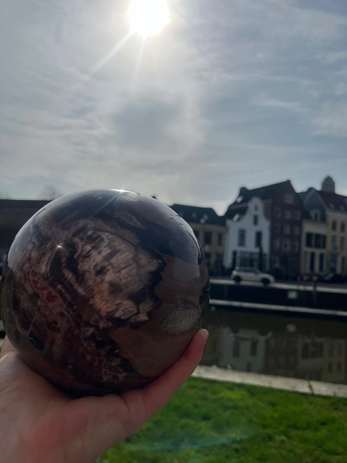 Een hand houdt een glanzende, ronde steen of bol voor zich. Op de achtergrond zijn kleurrijke huizen en een waterkant zichtbaar, onder een bewolkte hemel met de zon die schijnt.