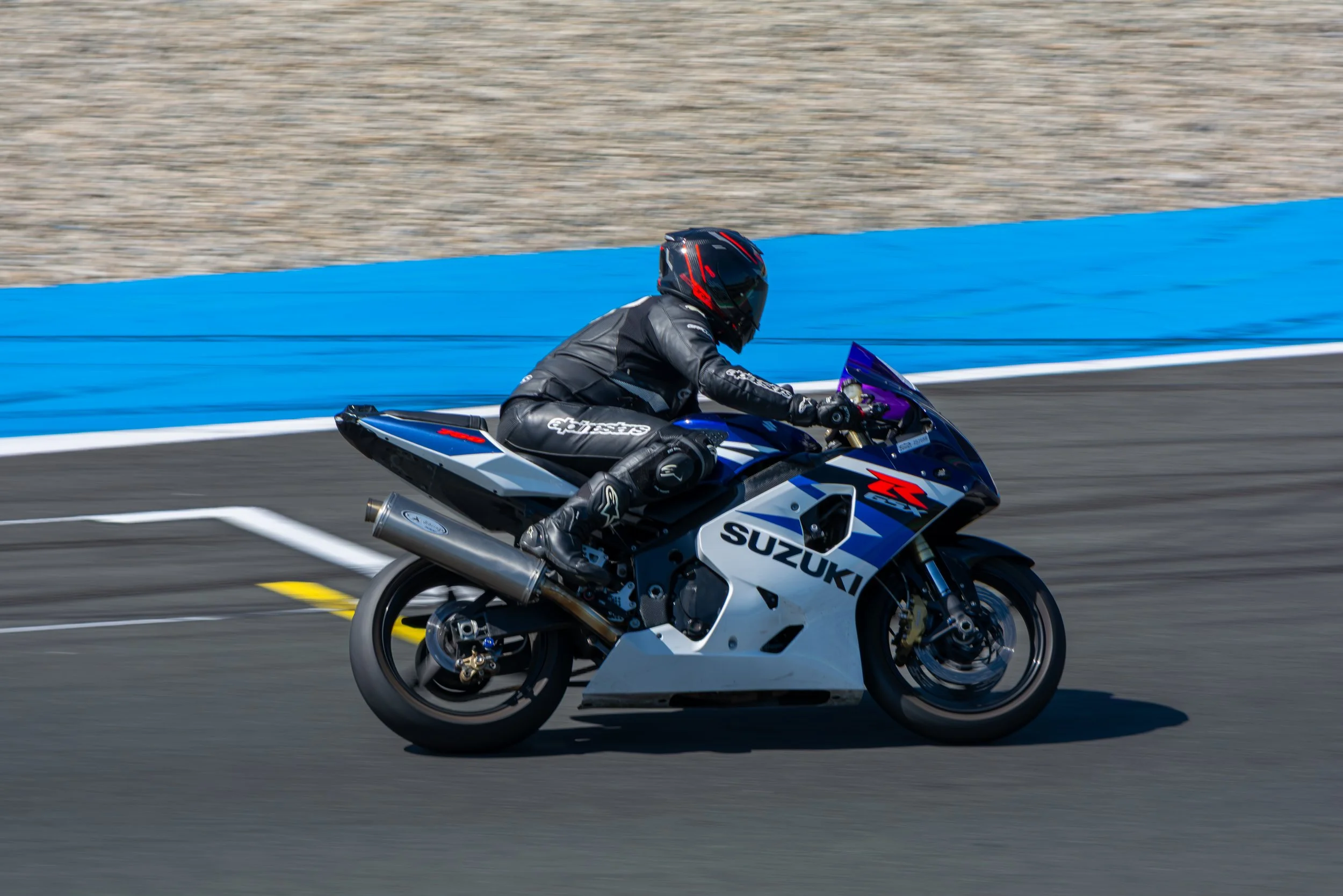 Een motorrijder in een zwart helm en zwart pak rijdt op een Suzuki sportmotor op een racebaan met blauwe vangrails.
