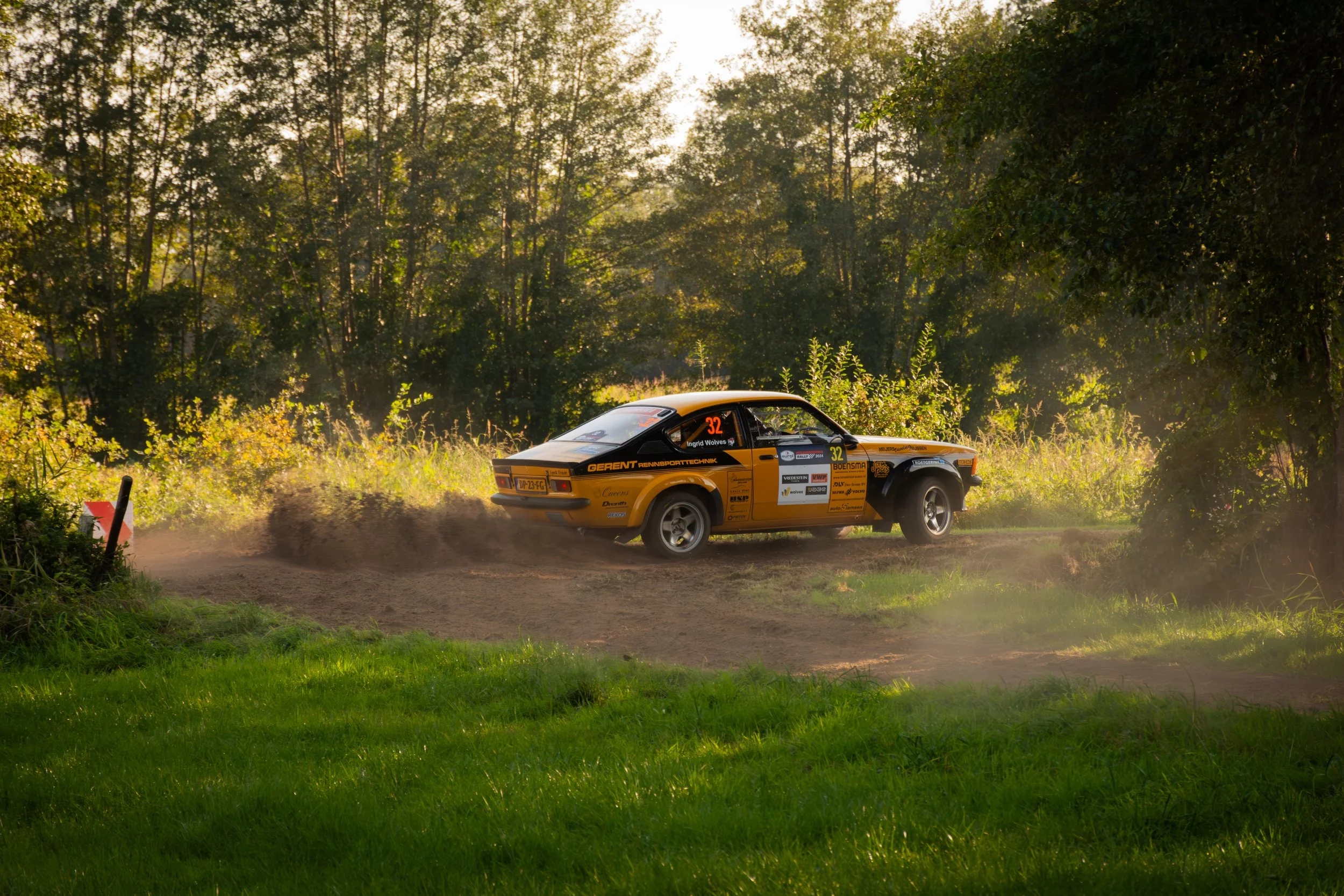Een geel Porsche rallyauto rijdt over een modderpad door een bos, stof en modder vliegen op.