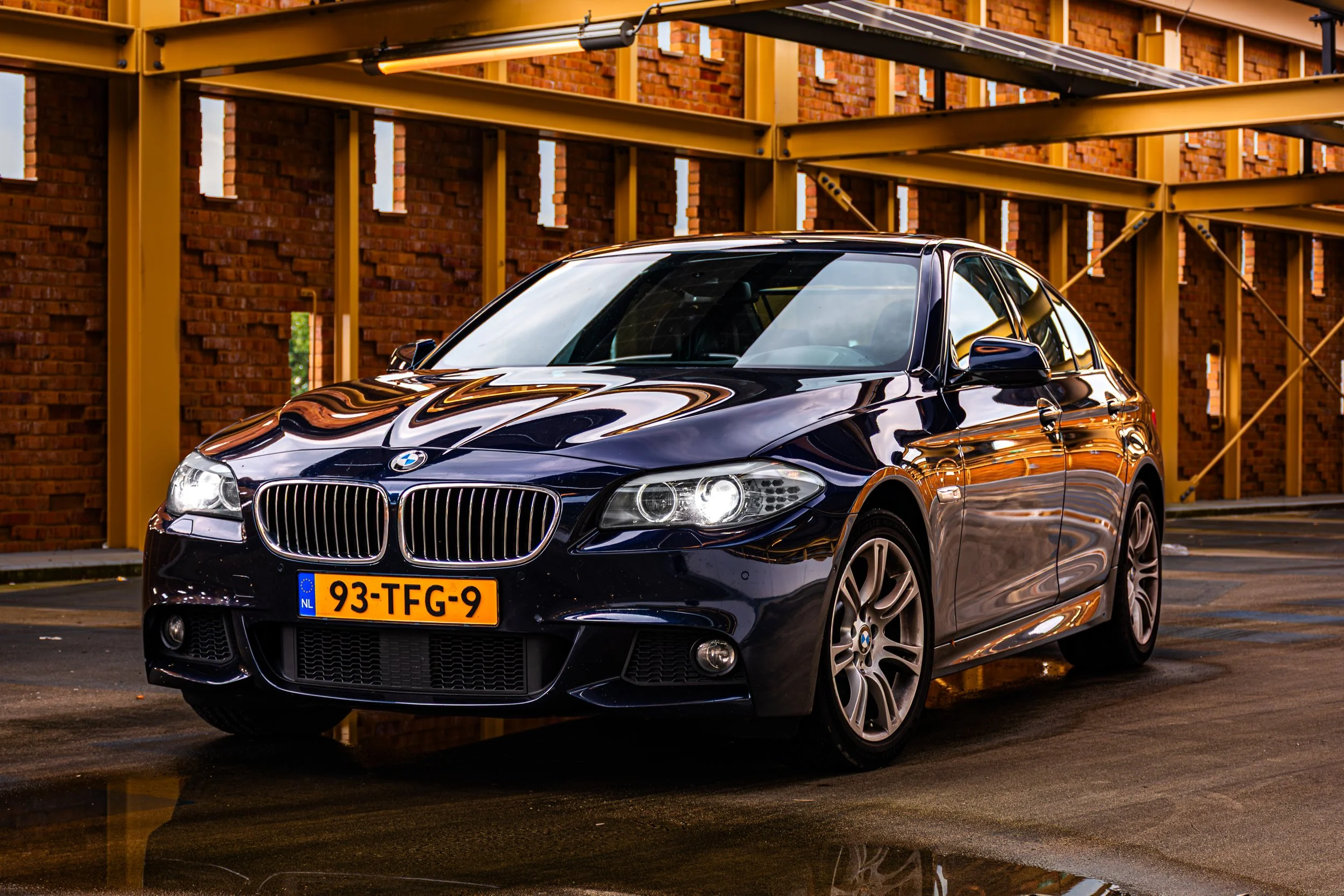 Zwarte BMW sedan met een Nederlands kenteken, geparkeerd voor een bouwplaats met gele steiger en een bakstenen muur.