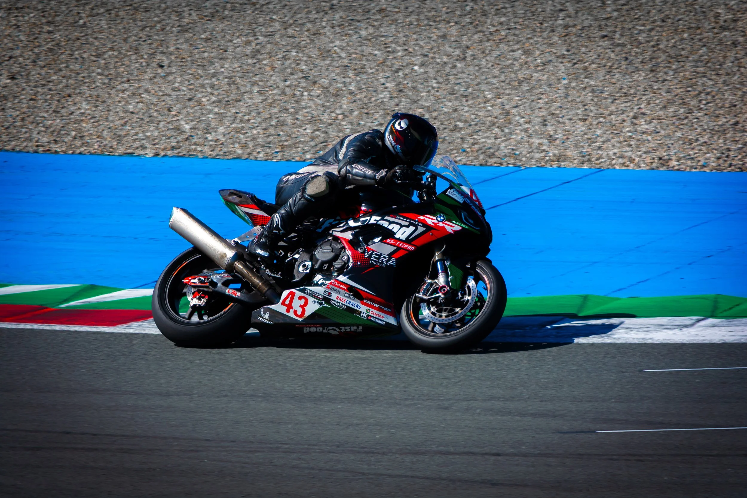 Een motorrijder in zwart pak en helm rijdt op een racebike met het nummer 43 op een circuit met groene, witte en rode markeringen en een asfaltbaan.