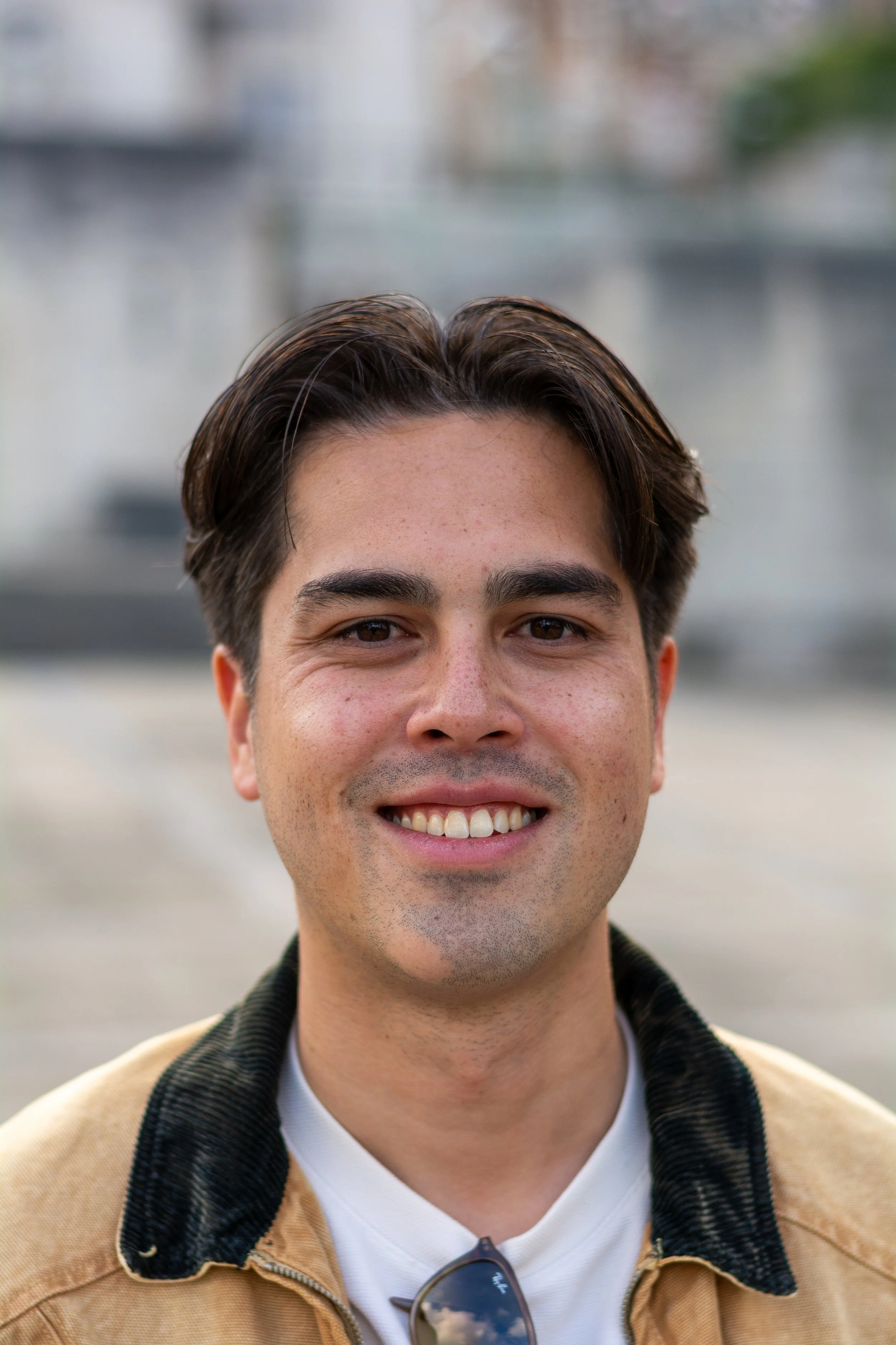 Close-up van een lachende man met donker haar, donkere ogen, in een buitenomgeving, gekleed in een beige jas en een witte shirt, met een paar zonnebrillen aan de hals.