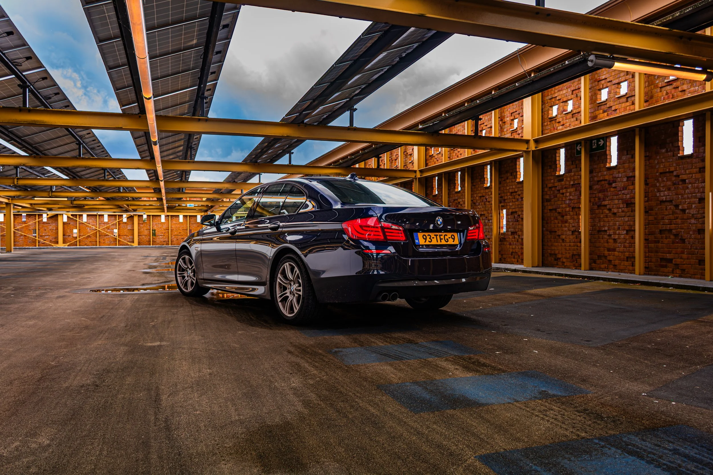 Een zwarte BMW-automobiel staat in een ondergrondse parkeergarage met een dak van zonnepanelen en een stenen muur met kleine raampjes.