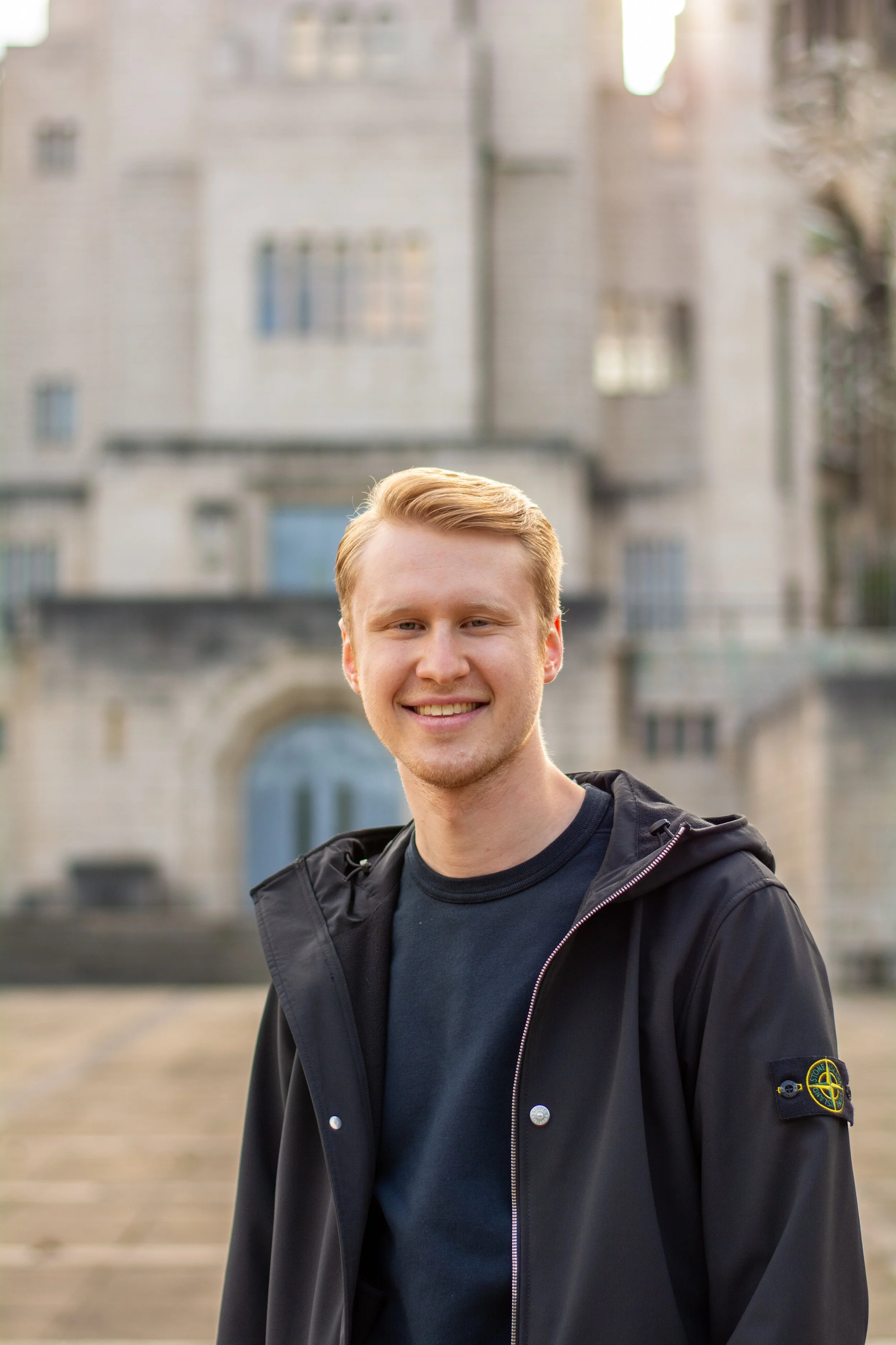 Een jonge man met blond haar, een zwarte jas en een blauwe shirt, glimlacht terwijl hij buiten staat met een oude gebouw op de achtergrond.