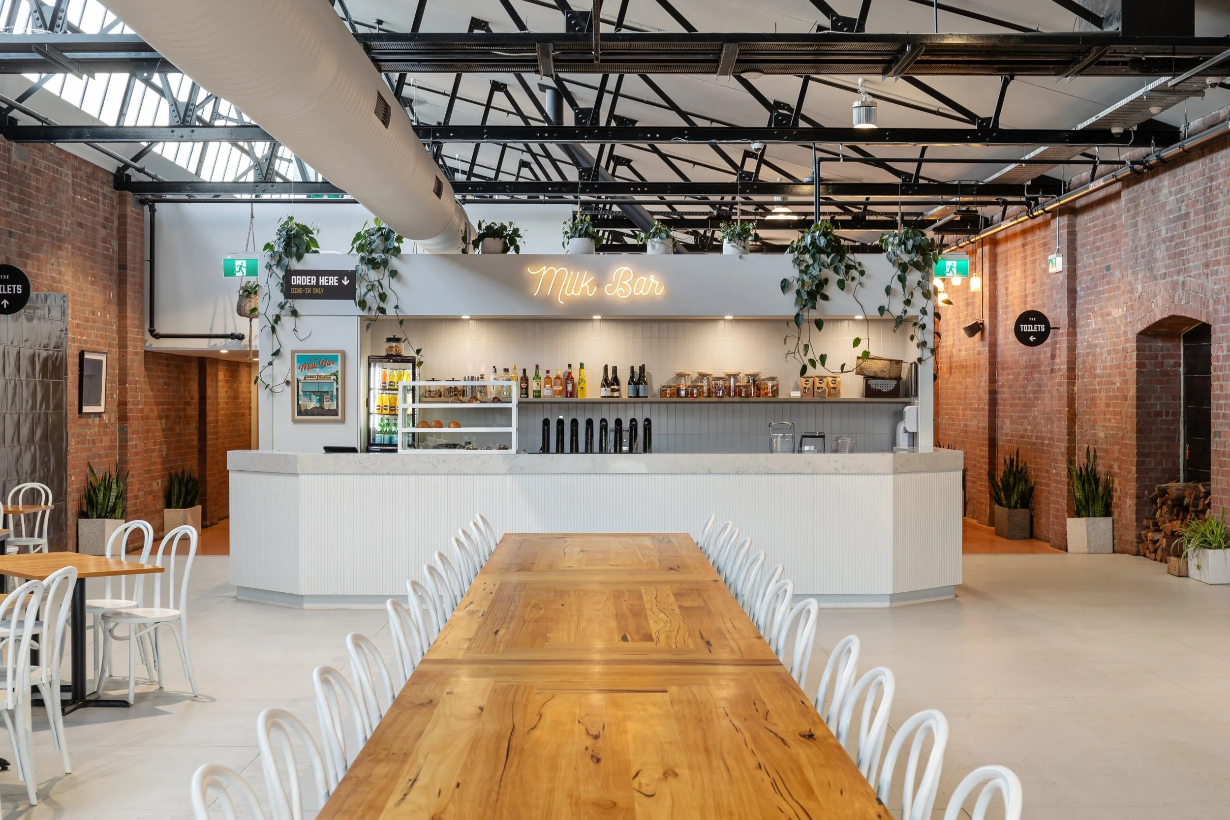 Interior of a modern cafe or restaurant with a milk bar counter, wooden tables and white chairs, exposed brick walls, ceiling pipes, hanging plants, and a neon sign that reads 'Milk Bar'.
