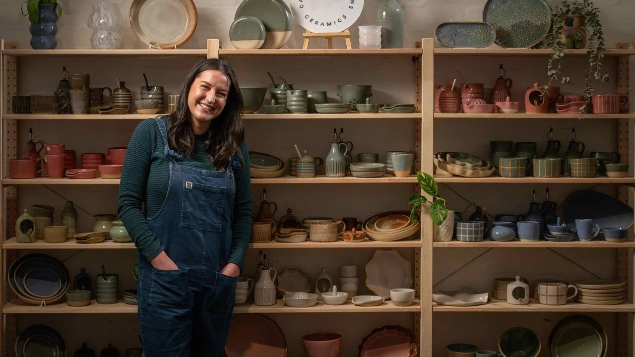 A smiling woman in a denim apron standing in front of wooden shelves filled with various ceramic dishes and pottery.