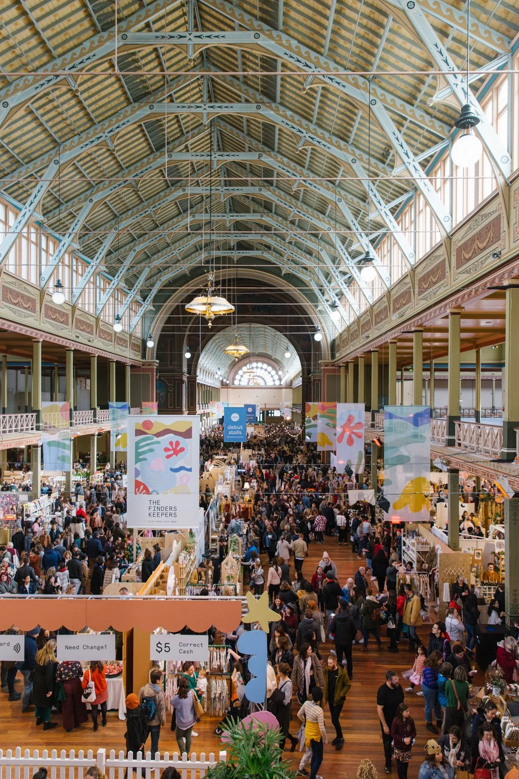 View of an indoor market or fair inside a large historic building with a high arched ceiling, filled with many people browsing stalls. Colorful banners and signs hang from the ceiling, and stalls are set up along the sides and center of the space.
