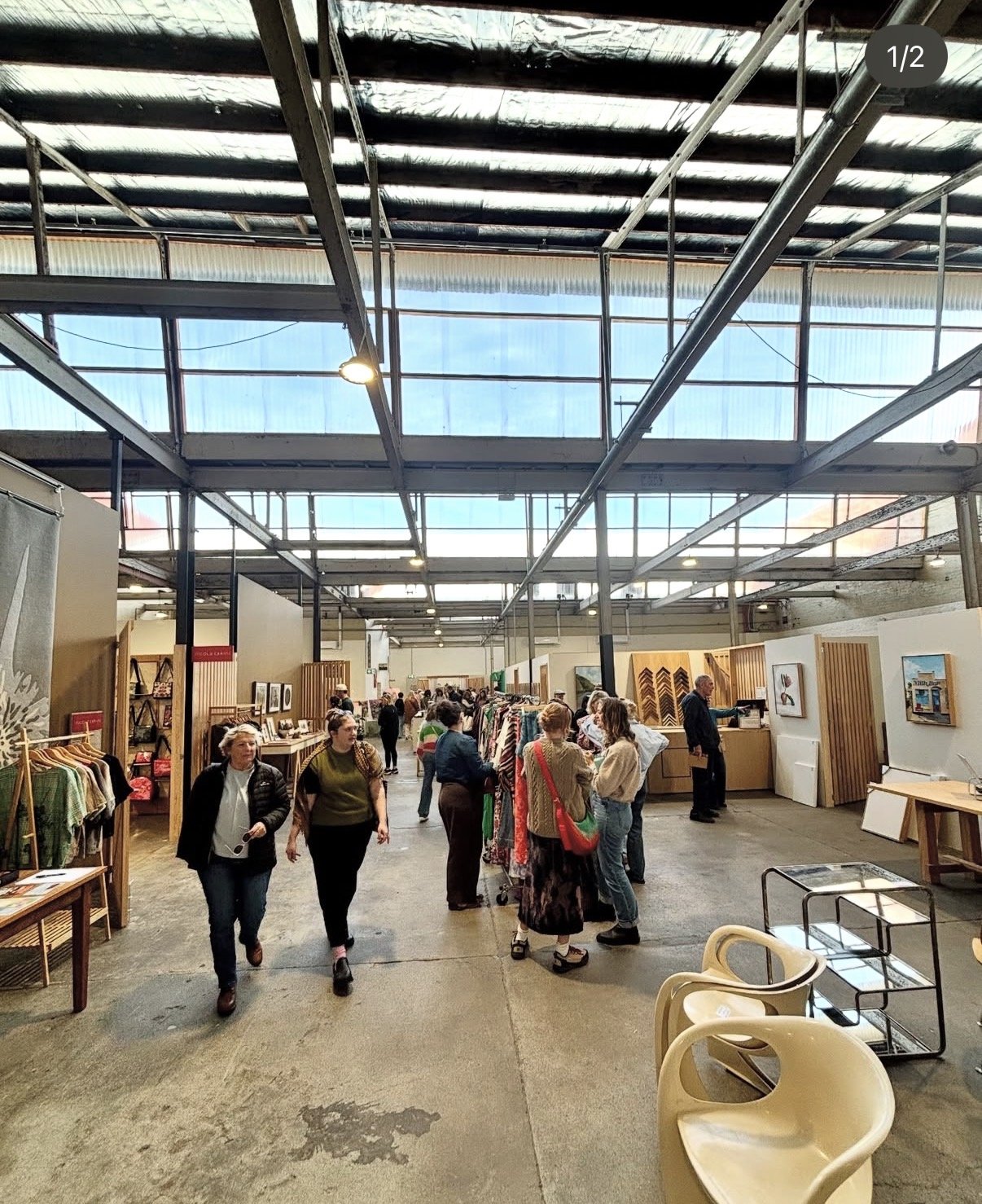 People browsing items at an indoor market or craft fair with clothing and artwork displays, in a spacious industrial-style building with high ceilings and metal beams.