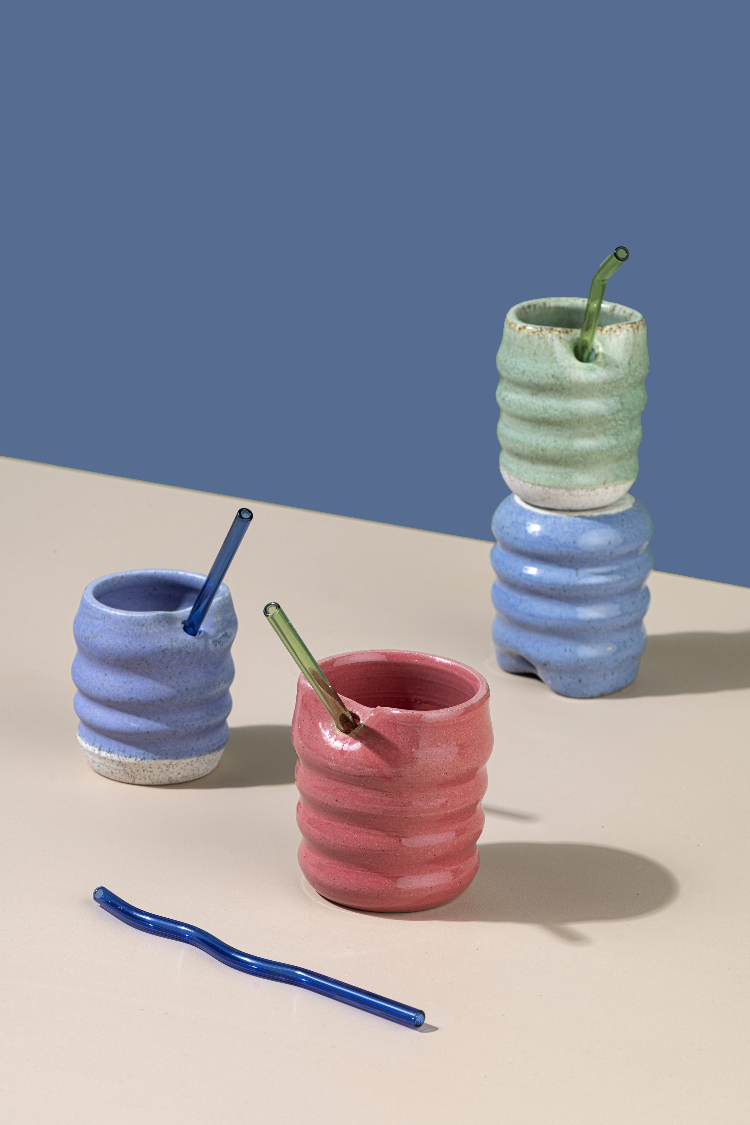 Colorful ceramic cups in pastel pink, blue, green, and red with striped patterns, some with bendy straws inside on a beige and blue background.