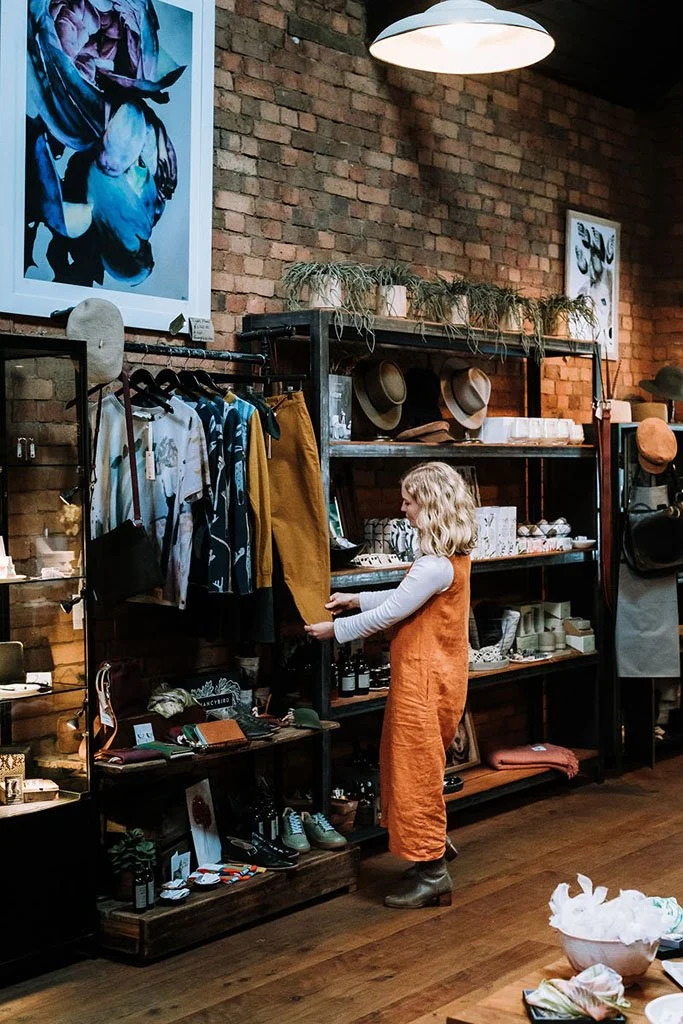 Woman with blonde curly hair shopping for clothes in a boutique store, browsing a clothing rack with pants and shirts. The store has a brick wall, artwork, and shelves with hats and accessories.