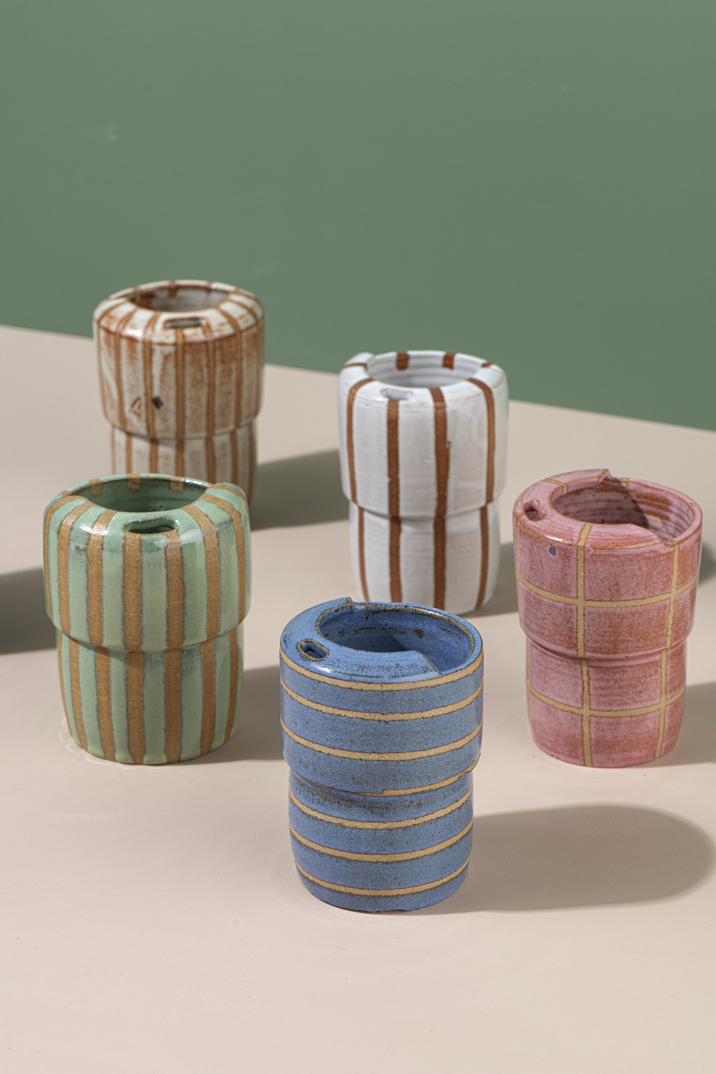 Colorful ceramic cups with vertical stripe patterns in pastel shades of orange, green, blue, and pink, arranged on a beige surface with a green background.