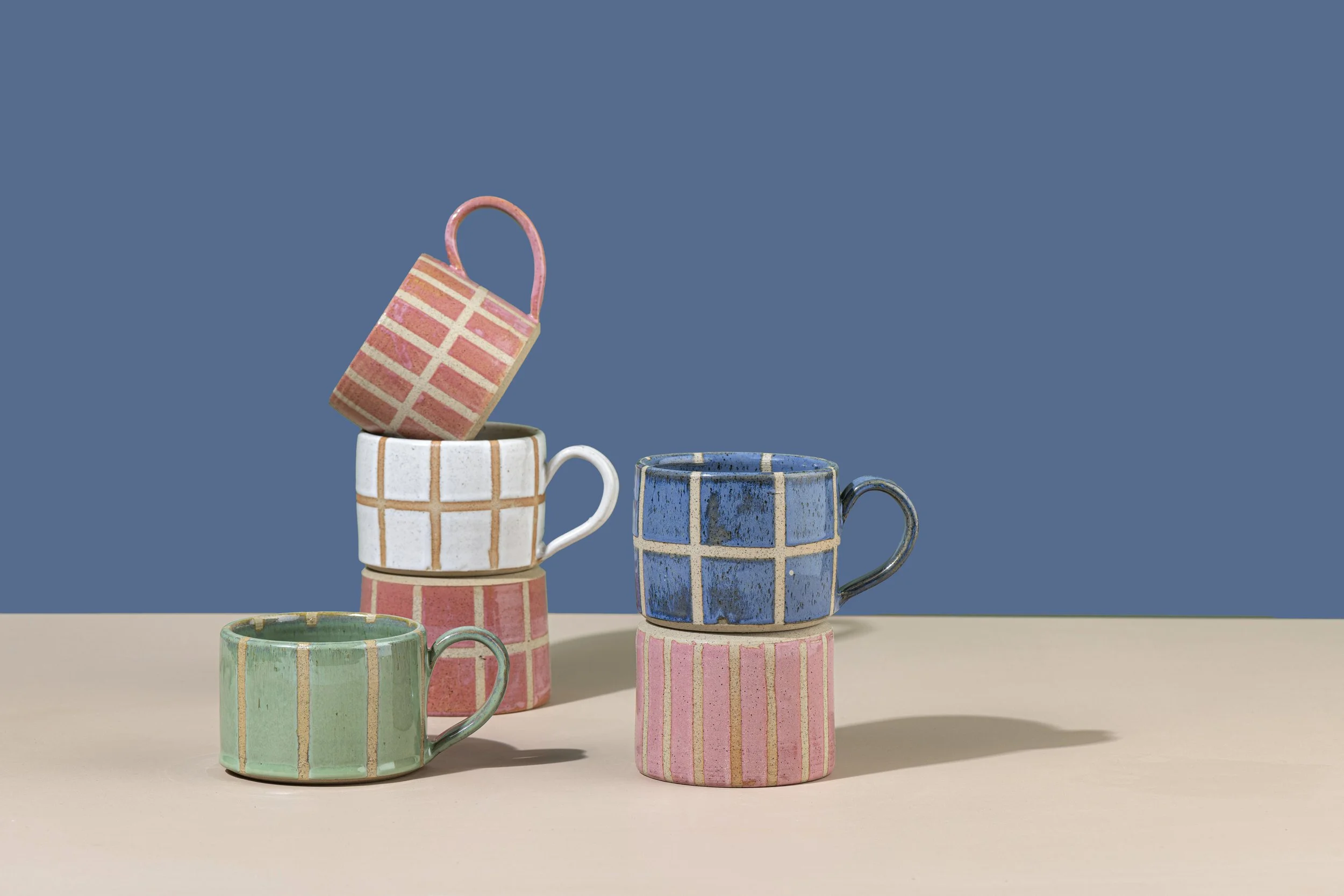 Colored ceramic mugs with geometric pattern and different pastel colors, stacked and arranged on a plain surface against a solid blue background.