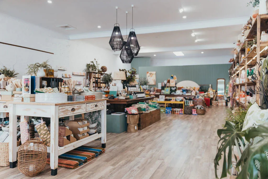 Interior of a bright boutique store with shelves and tables displaying various home decor, gifts, and accessories. The space features wooden flooring, modern black pendant lights, and a light-colored wall in the background.