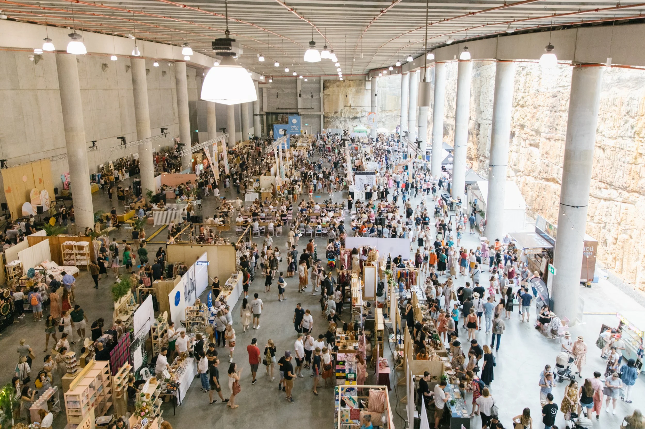 Inside a large indoor marketplace filled with stalls and booths, many people shopping and walking around, with high ceilings and large pillars, and rocky walls to the right.