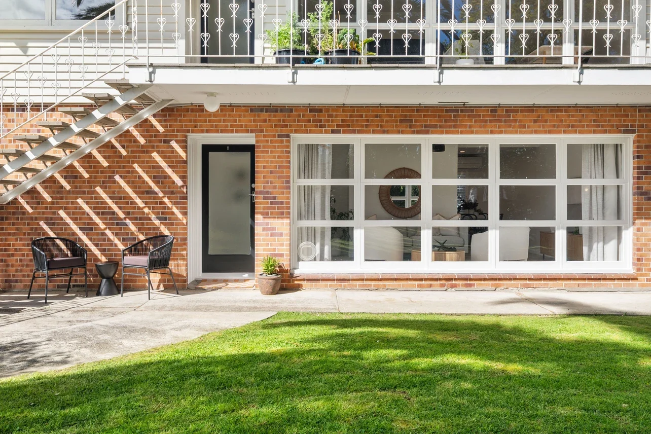 The image shows the exterior of a brick apartment building with a ground floor patio door and a large window. There are two black chairs with cushions and a small black side table on the patio, along with a potted plant near the door. An outdoor staircase leads to the upper floor, which has a balcony with railing and potted plants.