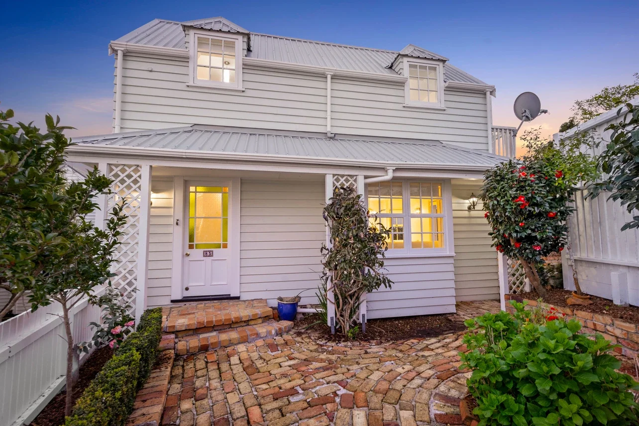 Front of a white house with a brick pathway, lit windows, plants, and a satellite dish on the side during sunset.