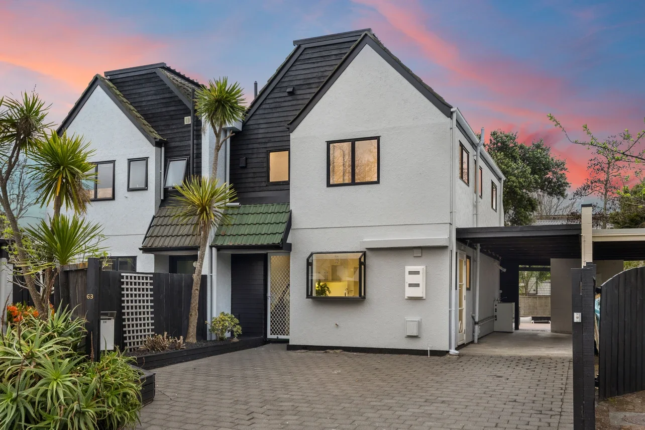 Modern multi-story house with white and black exterior, surrounded by plants and trees, during sunset with pink and purple sky.