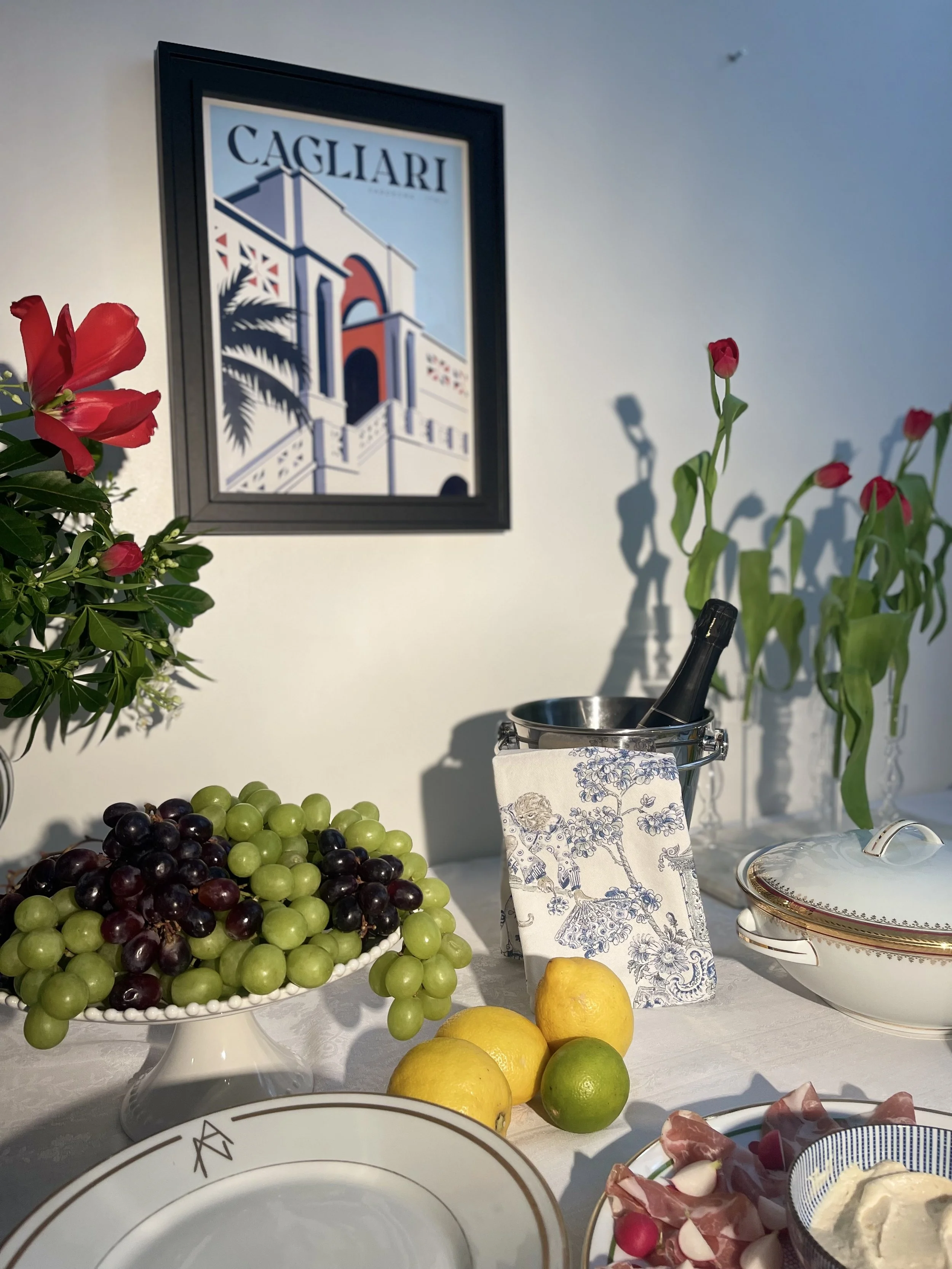 Une table garnie de fruits, fleurs, vaisselle et aliments pour un repas ou une dégustation, avec une décoration élégante et un tableau représentant Cagliari en arrière-plan.