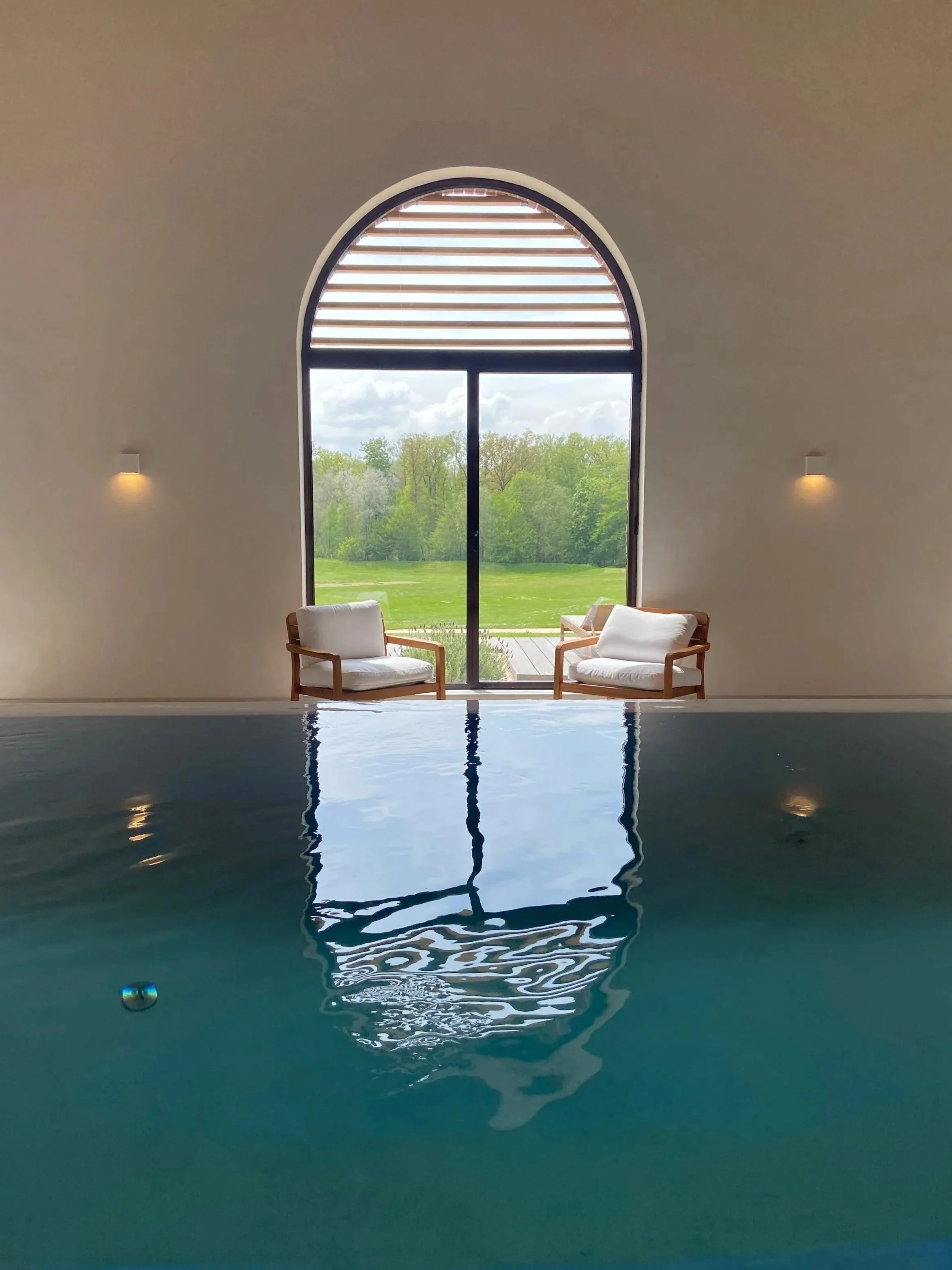 Piscine intérieure avec deux fauteuils près d'une grande fenêtre donnant sur un paysage verdoyant.