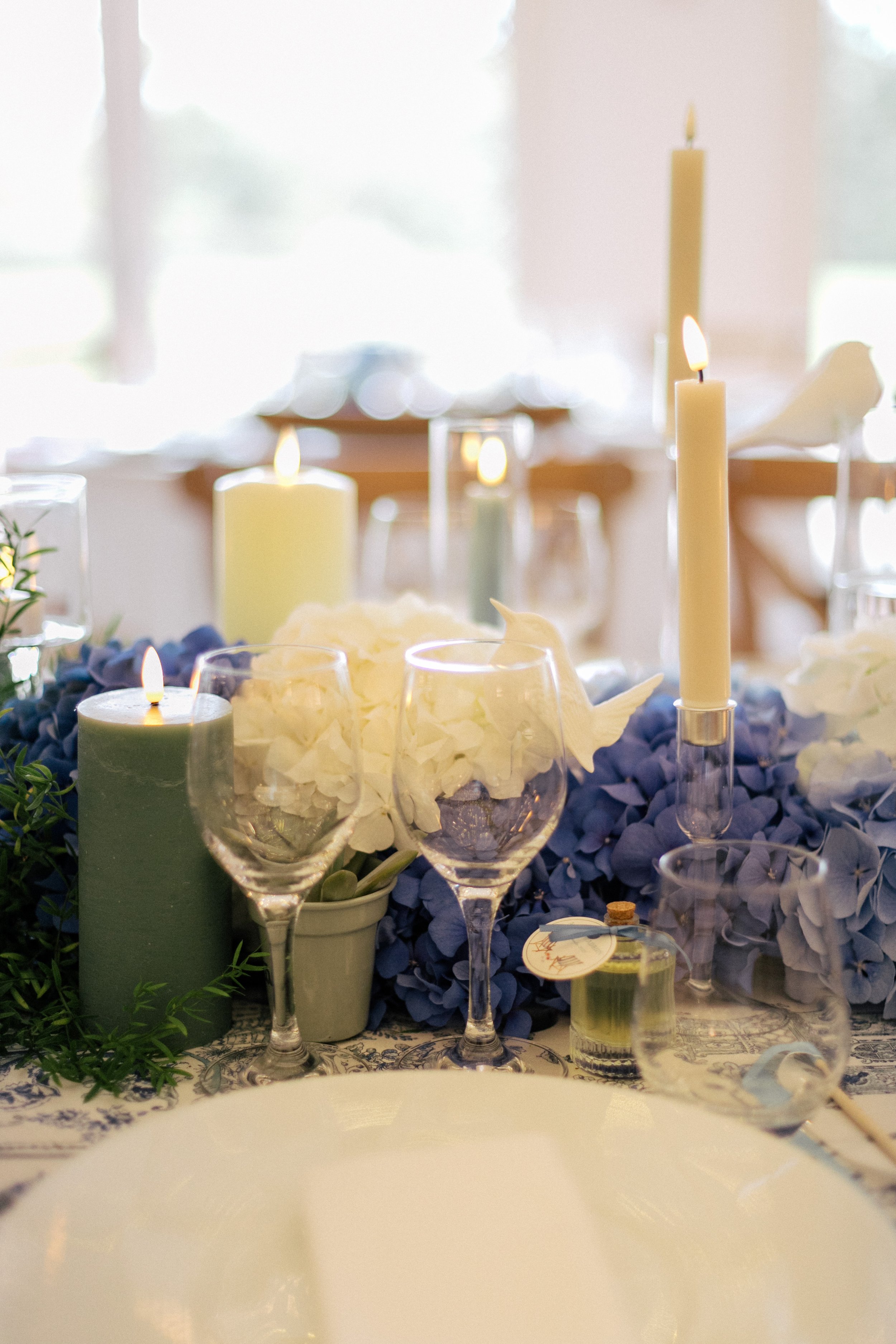 Décoration de table raffinée et élégante avec bougies blanches et vertes, fleurs bleues et blanches, et verre à vin, pour une occasion spéciale.