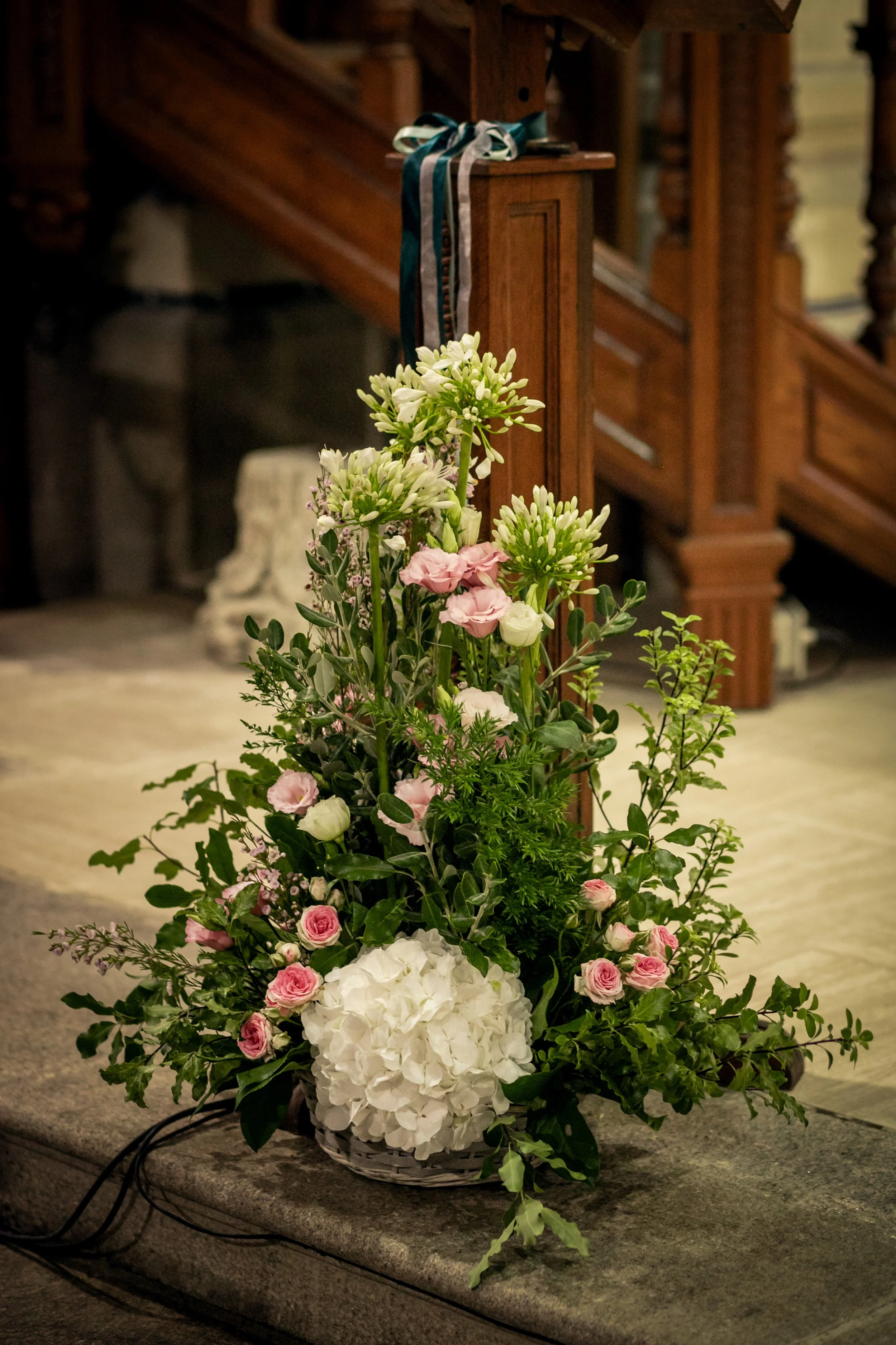 Un arrangement floral composé de roses, hortensias blanches, et autres fleurs, sur une table en pierre devant un escalier en bois dans un lieu intérieur.