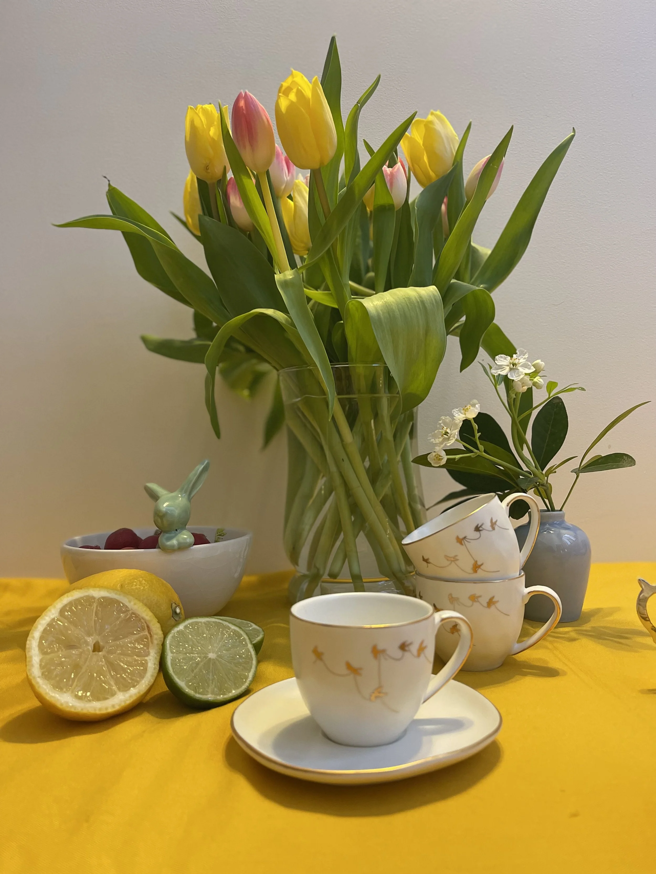 Un bouquet de tulipes jaunes et roses dans un vase transparent, des tranches de citrons et de concombres, plusieurs tasses en porcelaine avec un motif d'oiseaux, un petit lapin en céramique et un ramequin contenant des cerises rouges, tous disposés s