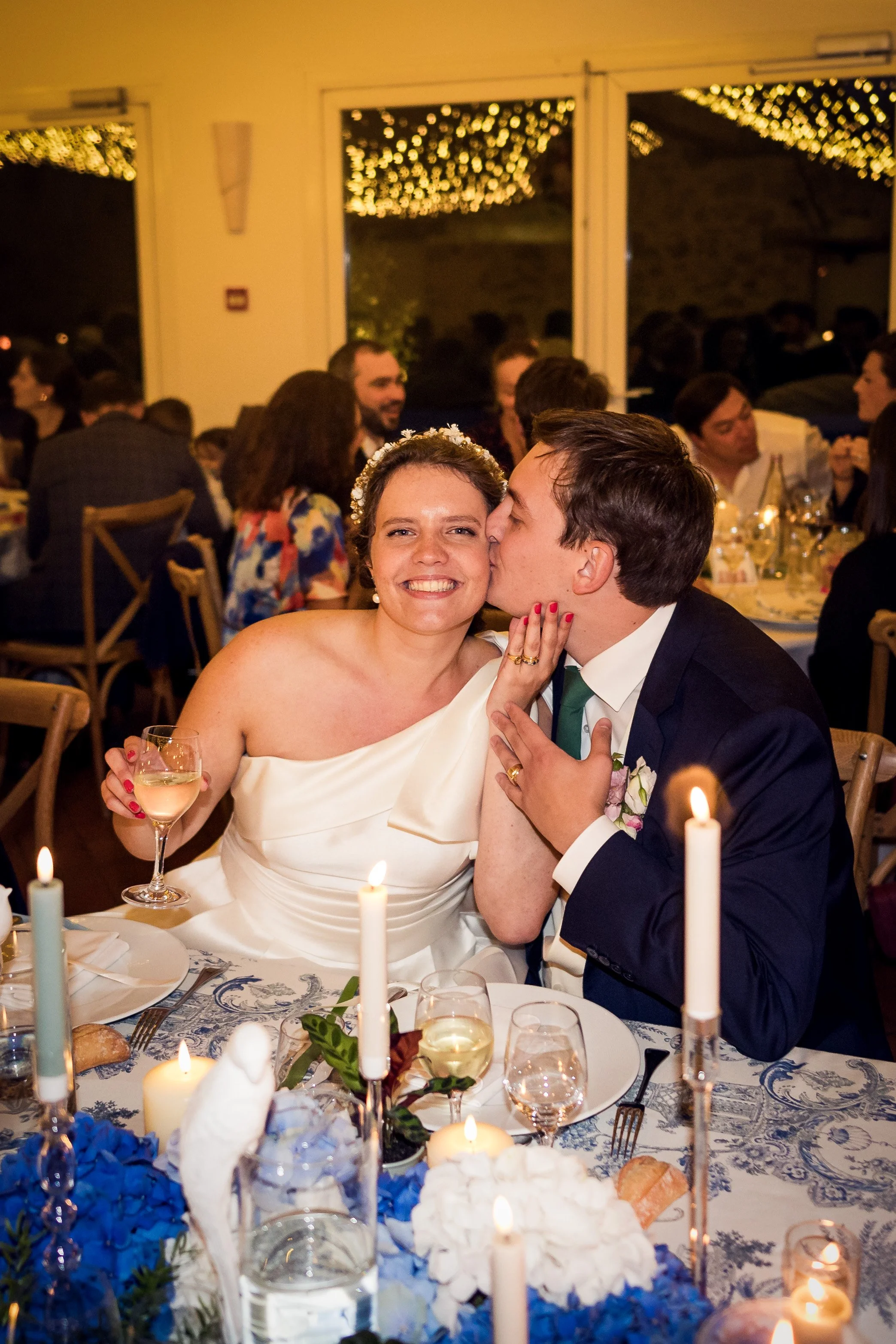 Un couple de jeunes mariés lors de leur mariage, la femme porte une robe blanche et un voile, le homme porte un costume. Ils sont assis à une table décorée avec des bougies et des fleurs, dans une salle de réception joliment éclairée.