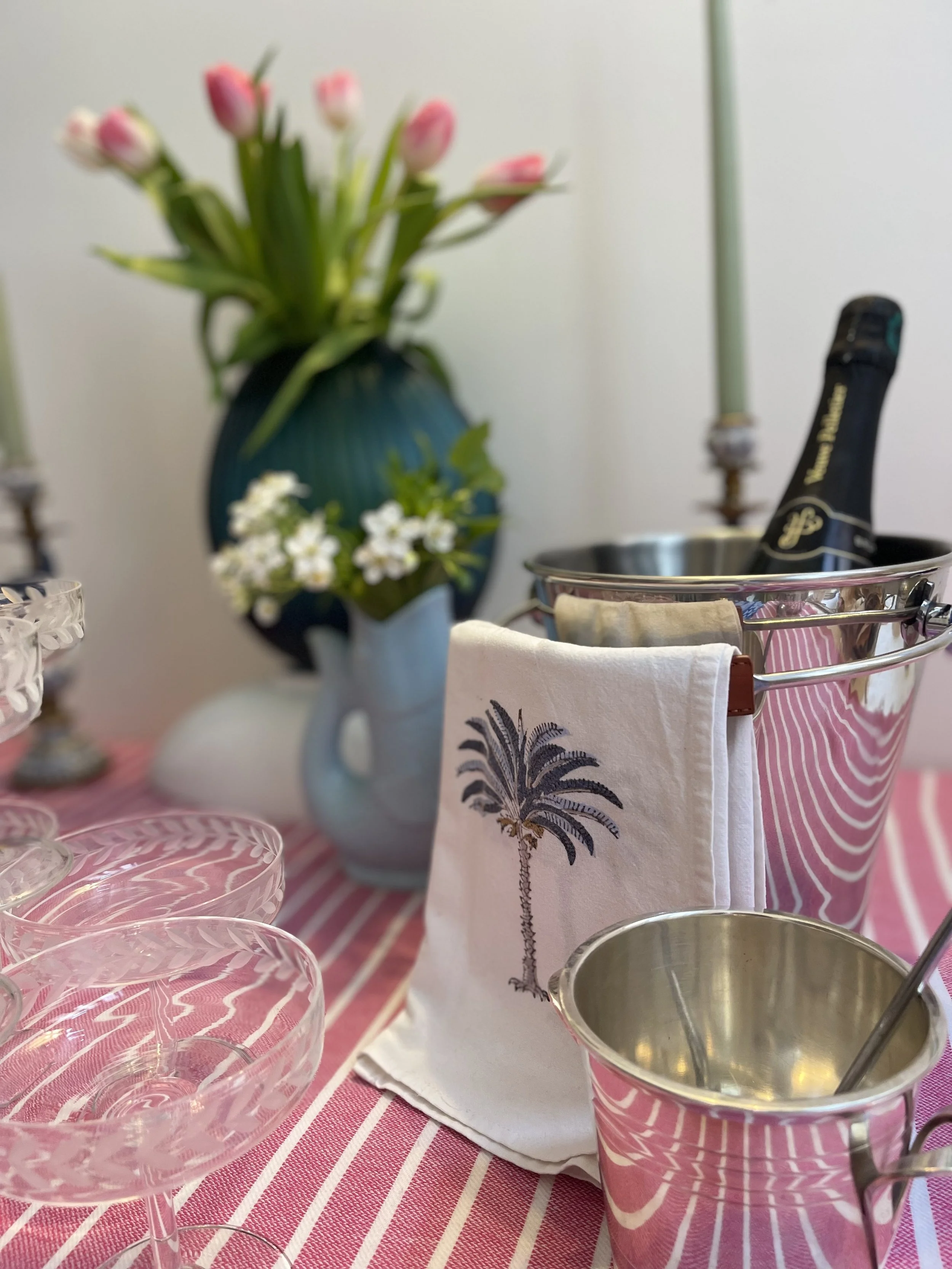 Décor de fête avec fleurs, vaisselle en verre, un seau à champagne avec une bouteille, et un plateau en métal, sur une nappe rose rayée.