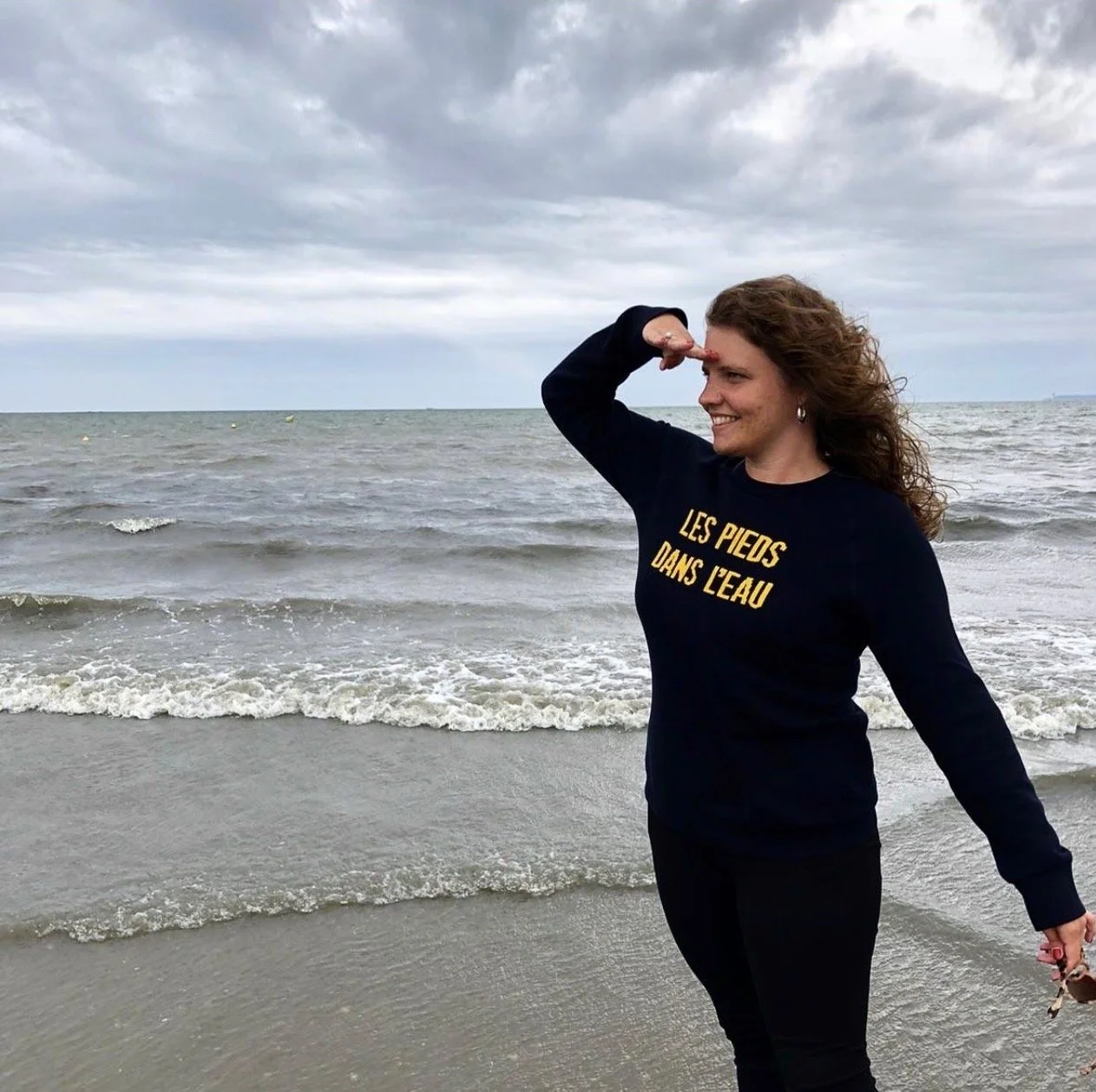 Une femme souriante sur la plage, portant un sweat noir avec l'inscription 'Les pieds dans l'eau', saluant ou saluant la mer avec la main en l'air, sous un ciel nuageux.