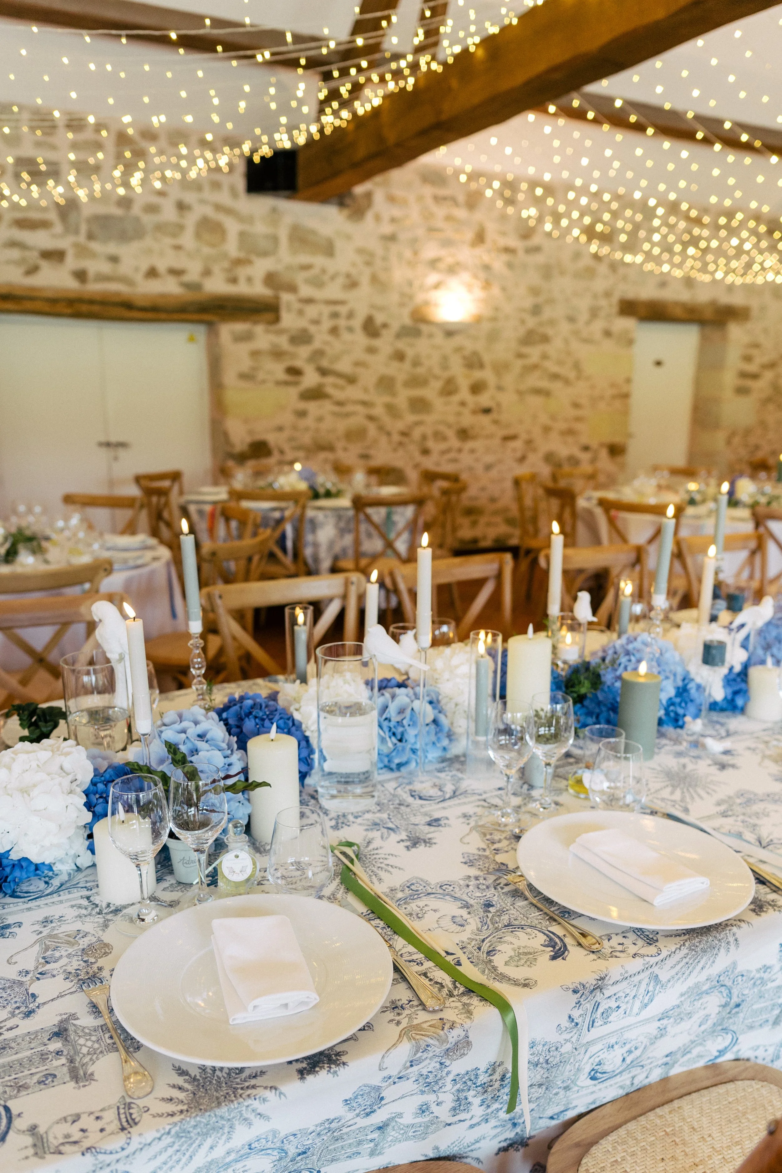 Une table décorée pour un événement, avec des draps en tissu blanc et une nappe à motifs bleus. La table est ornée de bougies, de fleurs bleues et blanches, de verres à vin et d'assiettes avec des couverts en or. En arrière-plan, il y a des chaises e