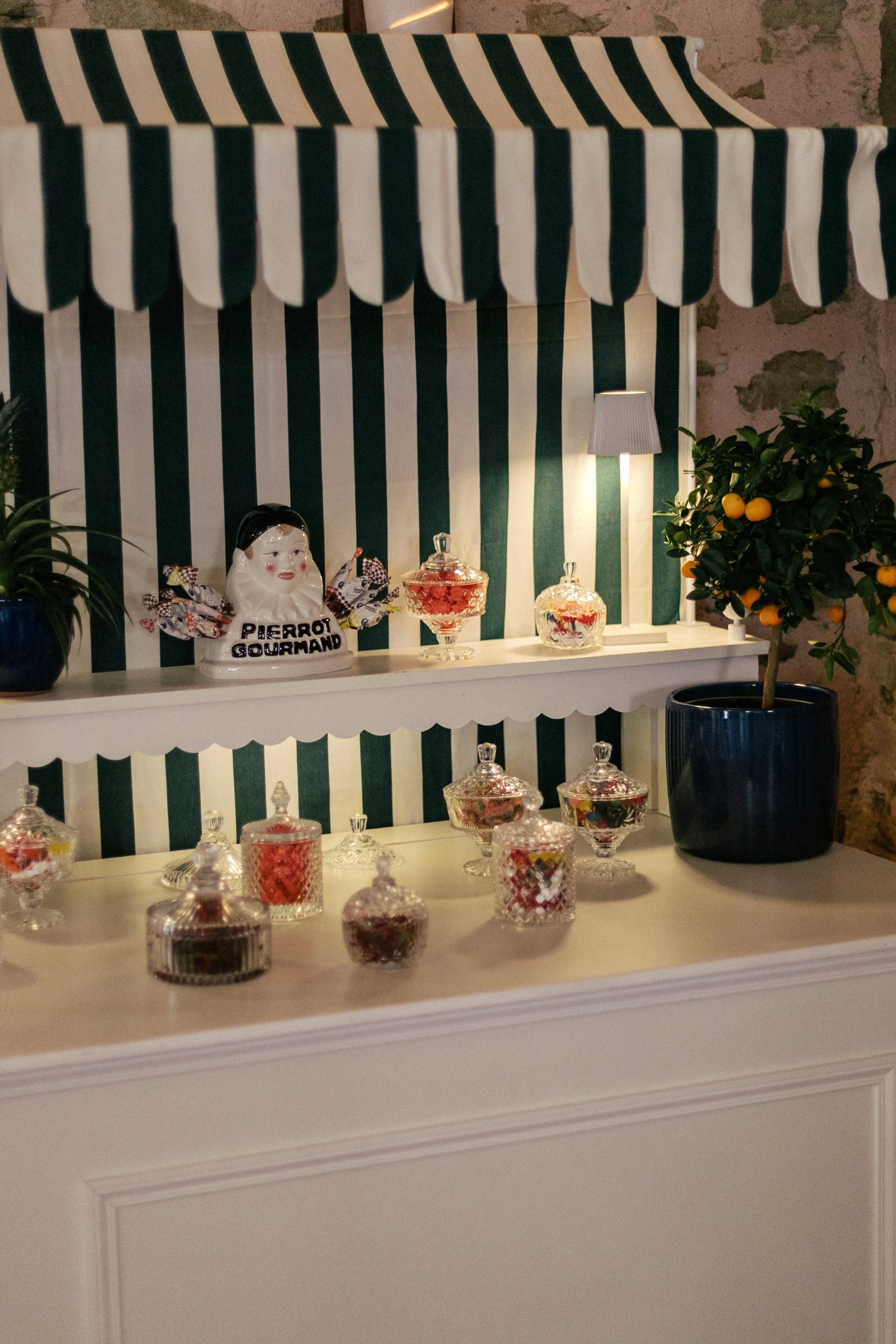 Un kiosque décorée avec des bocaux en verre, une figurine de Pierrot gourmand, une petite lampe, des agrumes en pot, et un mur en pierre en arrière-plan.