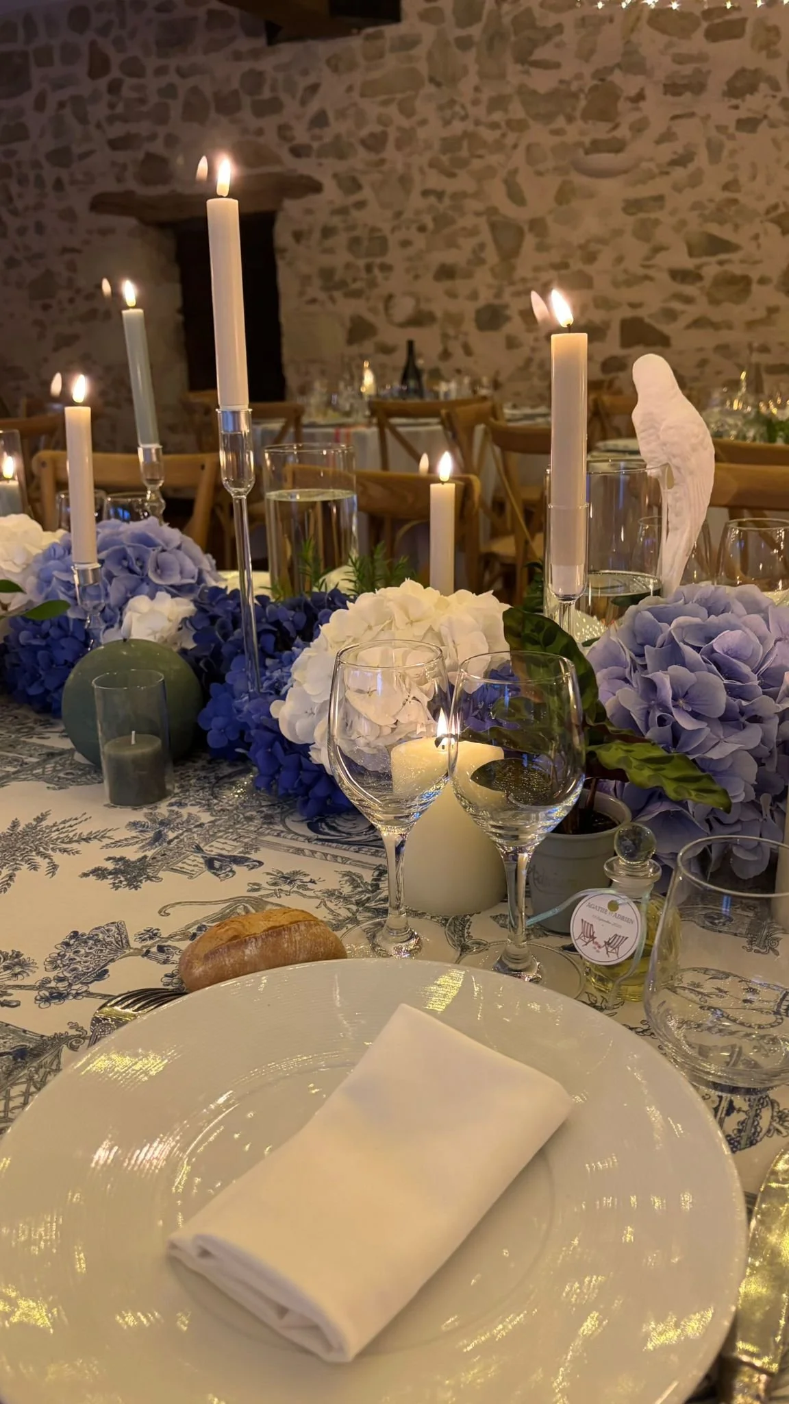 Table dressée pour un repas, avec des bougies allumées, des fleurs violettes et blanches, des verres, une assiette avec une serviette pliée et un croissant. Ambiance chaleureuse et élégante, mur en pierre en arrière-plan.