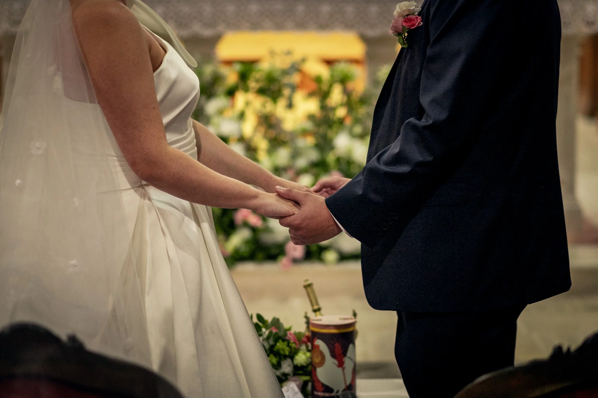 Un couple lors d'un mariage, ils se tiennent la main lors de la cérémonie. La femme porte une robe de mariée blanche avec un voile, et l'homme porte un costume noir avec une boutonnière.
