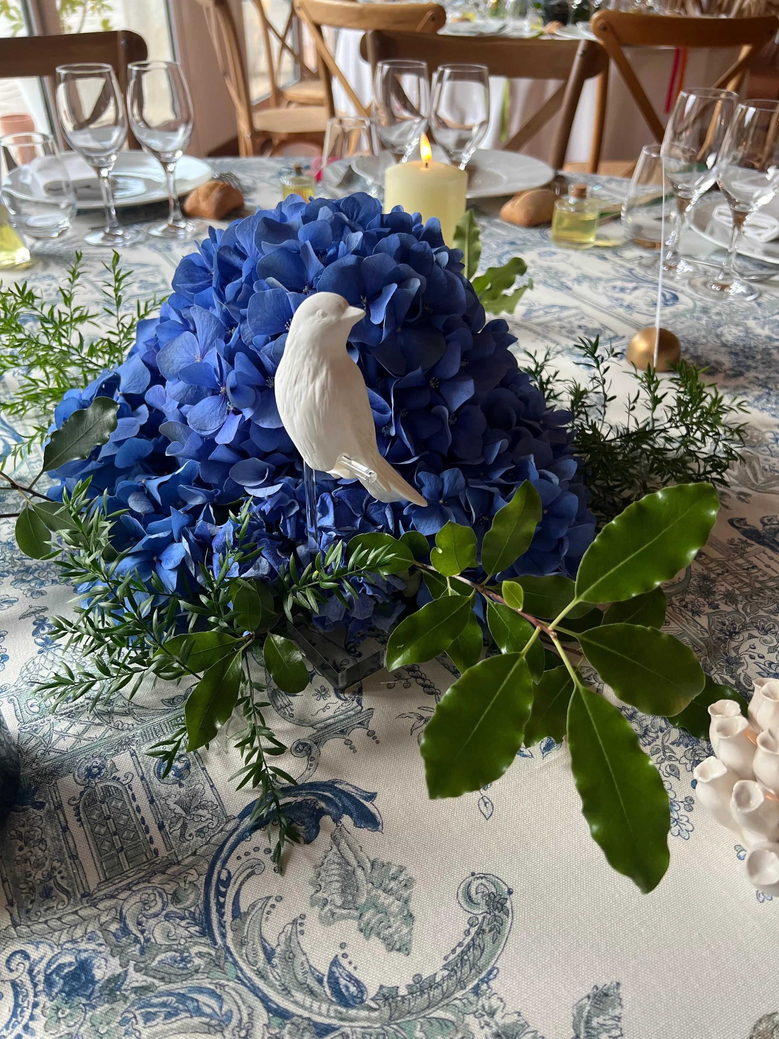 Une table décorée pour un repas, avec un centre de fleurs composé d'hortensias bleus, une petite statue blanche en forme d'oiseau, des branches vertes et des boules dorées. La table est recouverte d'une nappe à motif bleu et blanc, avec des verres, d