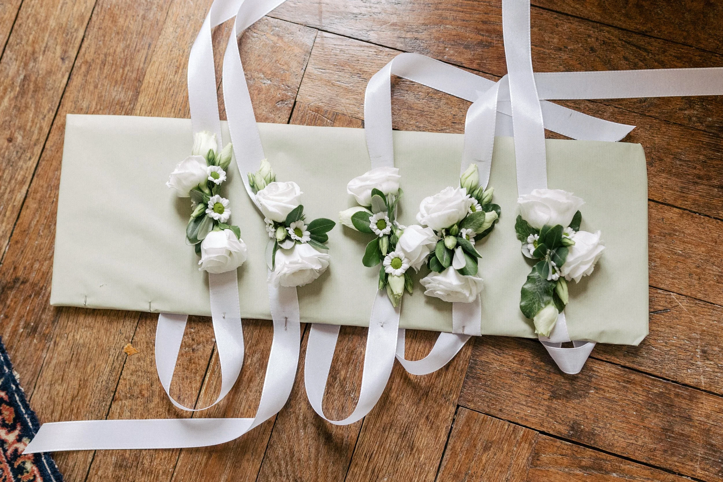 Un panier blanc décoré de fleurs blanches et de rubans satinés blancs, posé sur un plancher en bois. Ce sont des cadeaux pour les témoins de la mariées, le détail qui sublimera leurs tenues