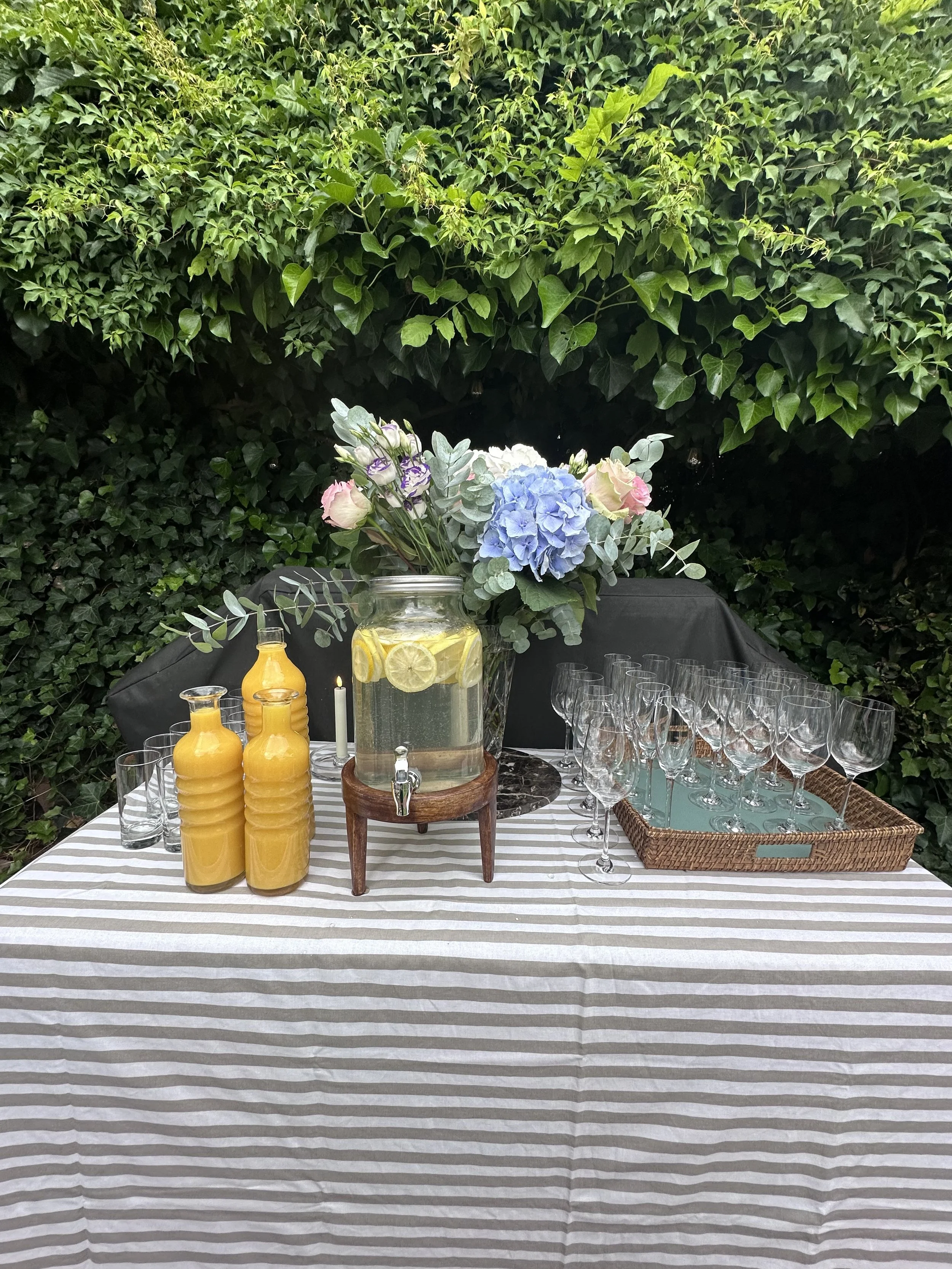 Une table decorée avec un vase de fleurs, des bouteilles de jus d'orange, des verres à vin vides, et un décor de jardin en arrière-plan avec des plantes vertes.