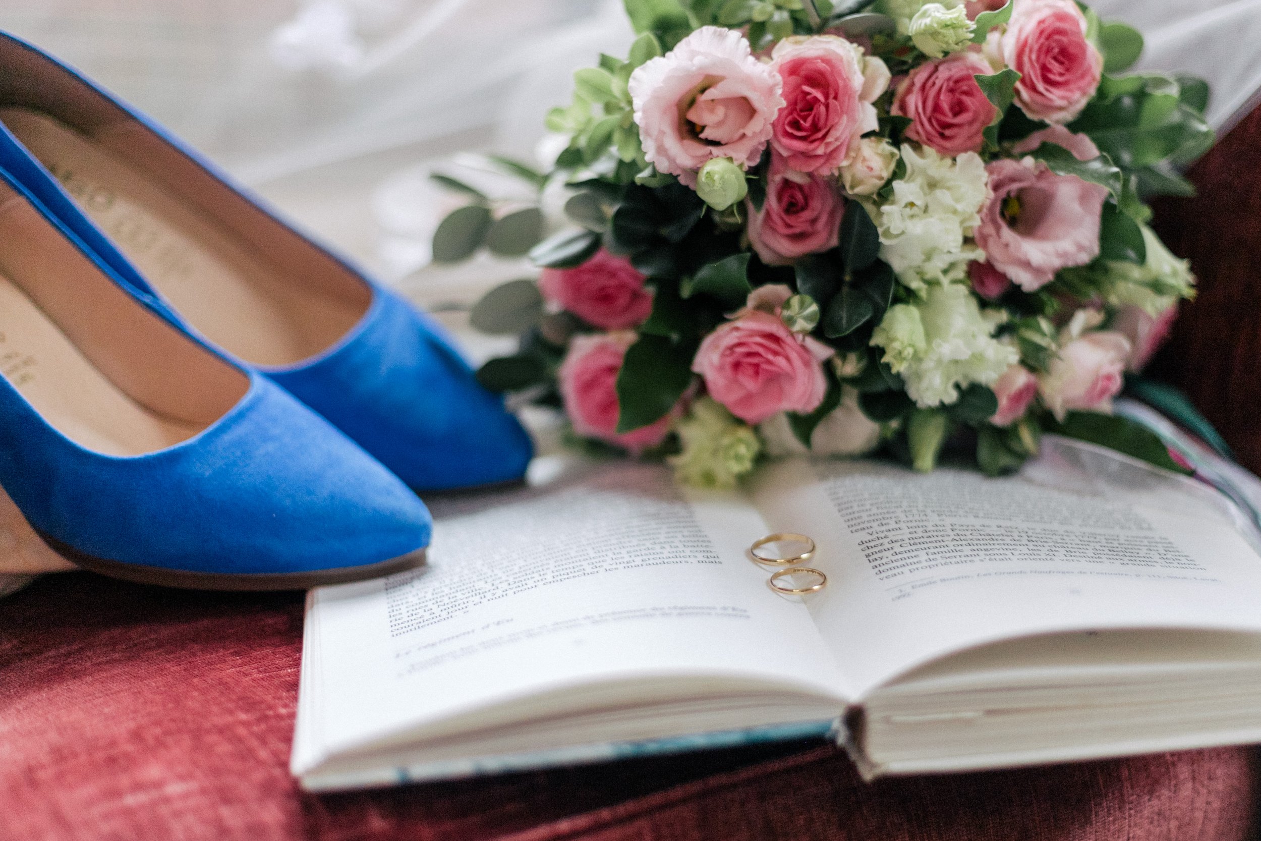 Une paire de chaussures bleues, un bouquet de fleurs roses et blanches, des alliances, un livre ouvert, pendant les préparatifs de la mariée.
