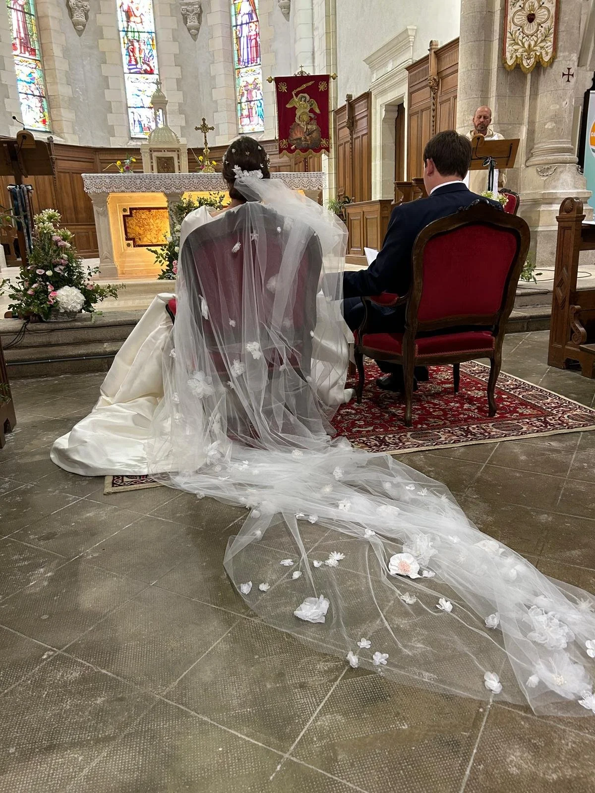 Un couple en mariage assis dans une église, lors de la cérémonie. La mariée porte une robe blanche avec un voile long et transparent décoré de fleurs blanches, et le marié porte un costume noir. L'autel est décoré de fleurs et le sol est en pierre. La scène se déroule à l'intérieur d'une église avec des vitraux colorés en arrière-plan.