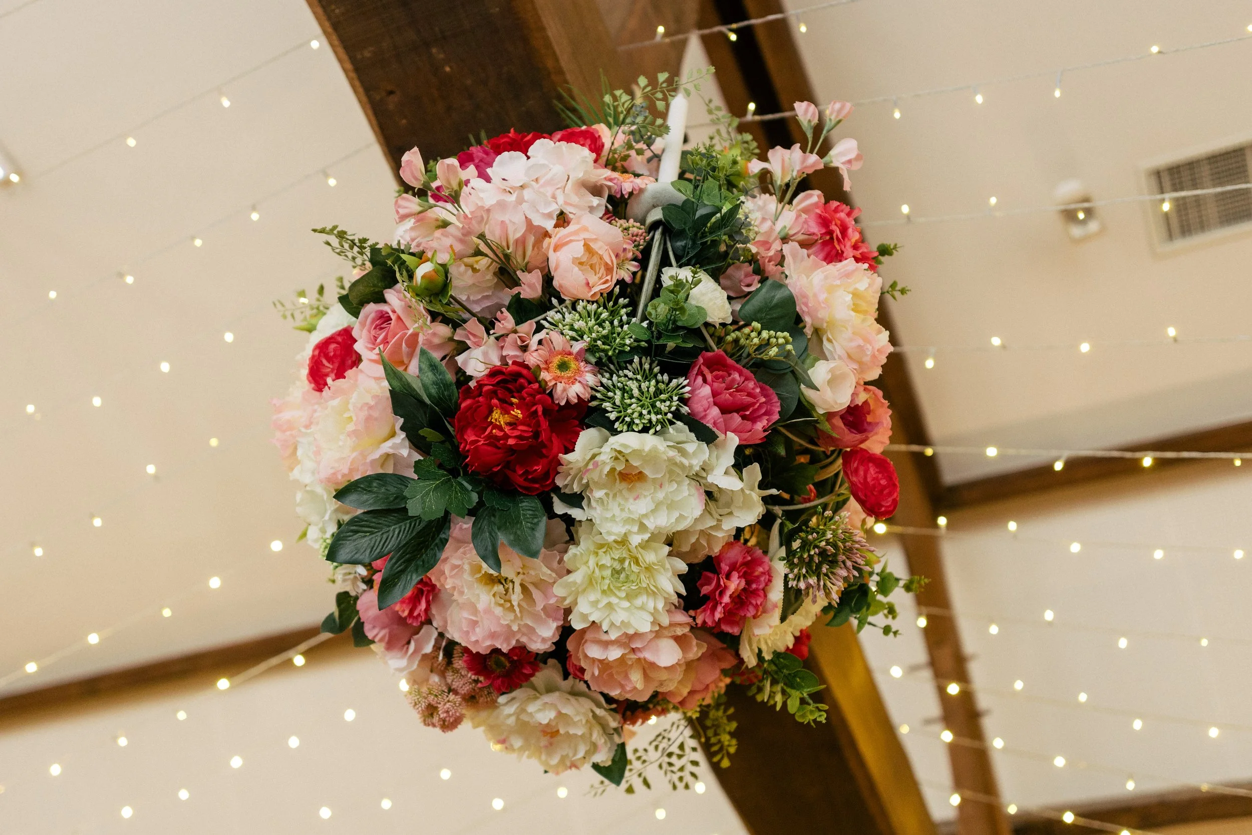 Un bouquet de fleurs roses, rouges et blanches suspendu au plafond, avec des guirlandes lumineuses en arrière-plan dans une salle de fête.