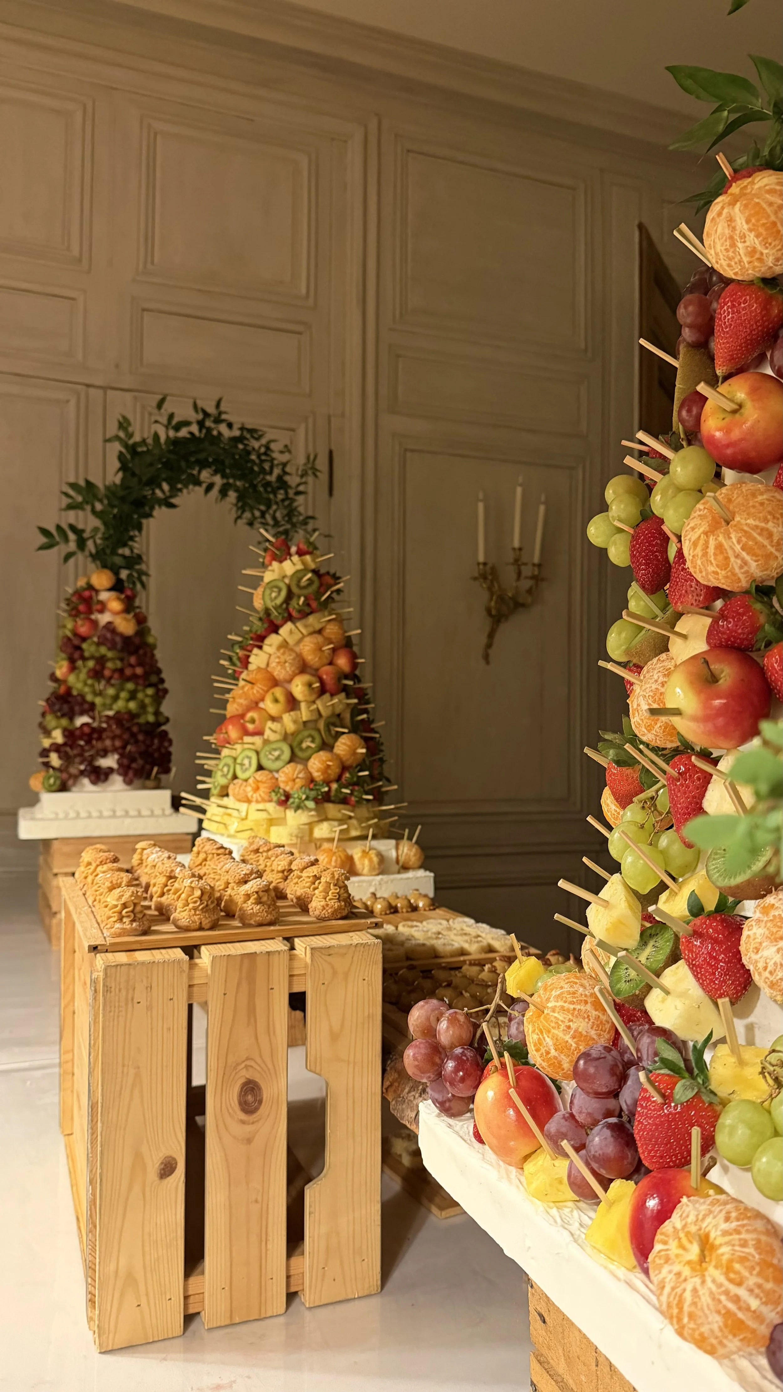 Présentation de buffet de fruits et pâtisseries dans une salle élégante, avec panneau en bois et chandelier ancien en arrière-plan.