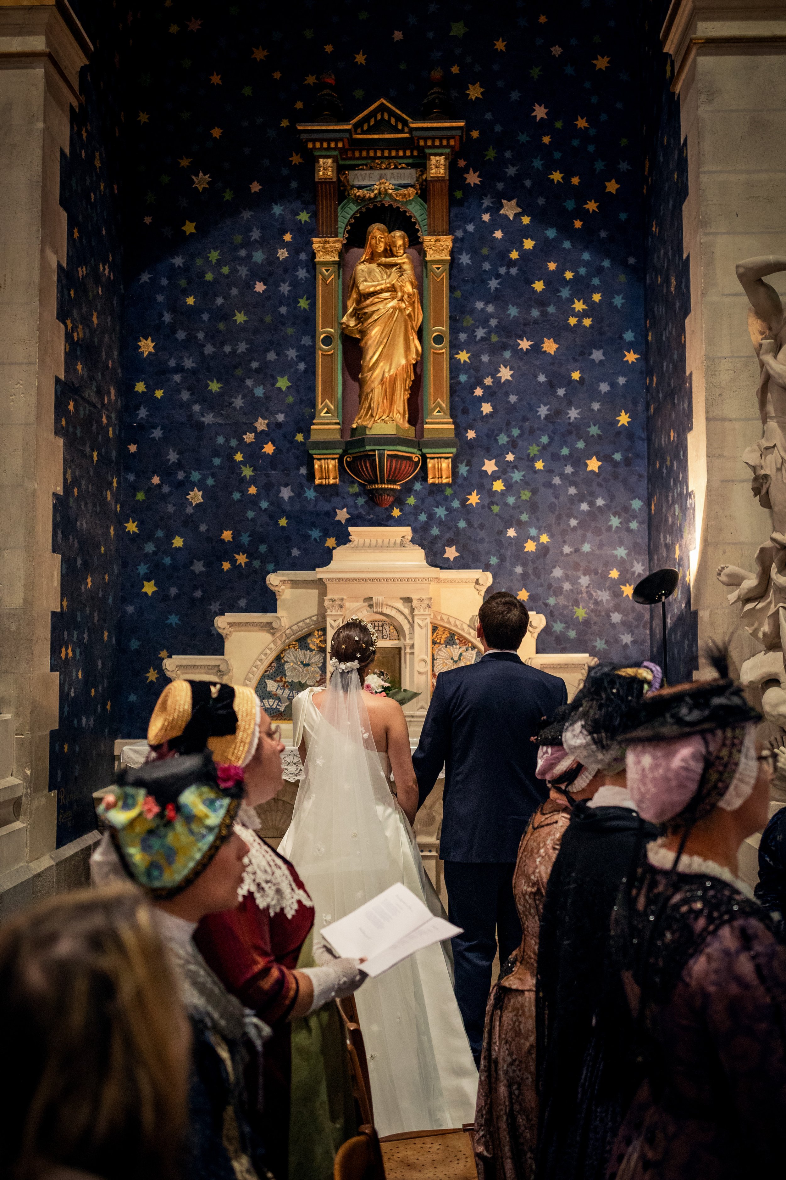 Une cérémonie religieuse dans une église avec un couple au mariage, entouré de personnes habillées en costumes et vêtements traditionnels. La scène se déroule devant une grande œuvre d'art représentant la Vierge Marie avec Jésus, dans un décor étoilé