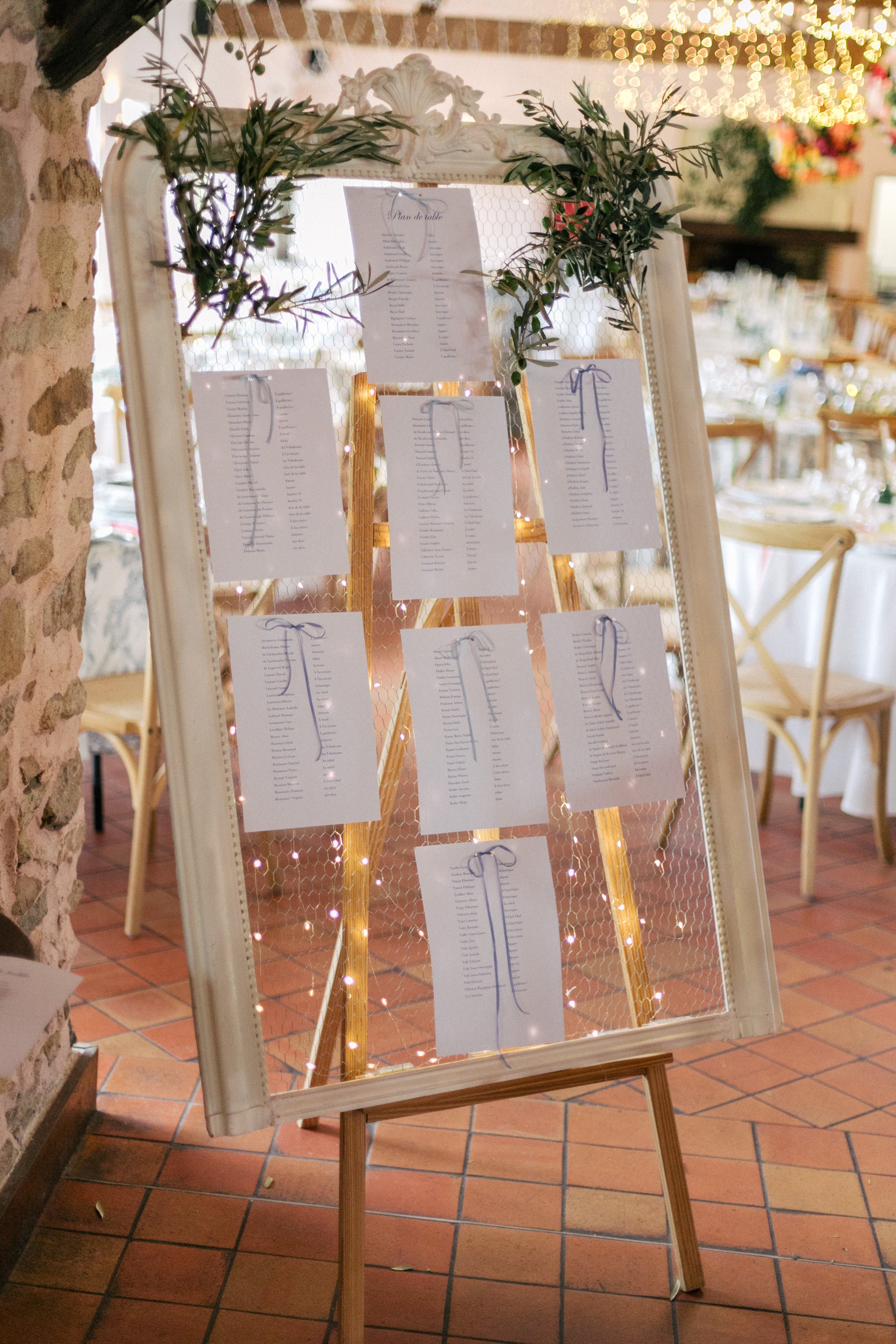Ardoise avec des plans de table de mariage décorée de branches d'olivier, entourée de lumières, dans une salle élégante avec tables et chaises en bois. Le cadre est celui d'un miroir ancien, chiné pour l'occasion. 