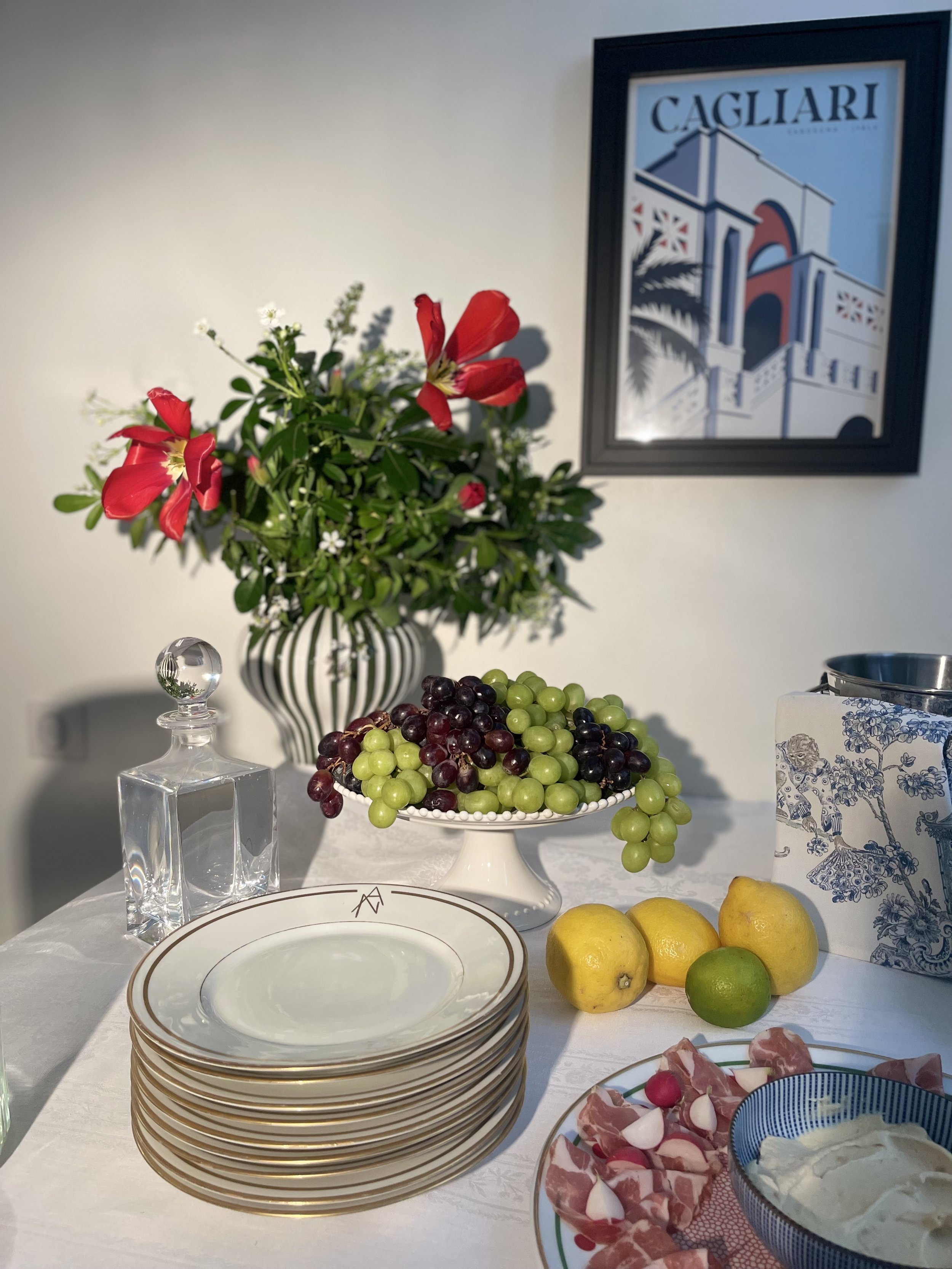 Une table dressée avec des assiettes, des fruits, une vase avec des fleurs, et un décor avec un tableau représentant un pont et une maison. On voit aussi des grappes de raisin, du citron et de la lime, ainsi que de la charcuterie, du fromage, et des 